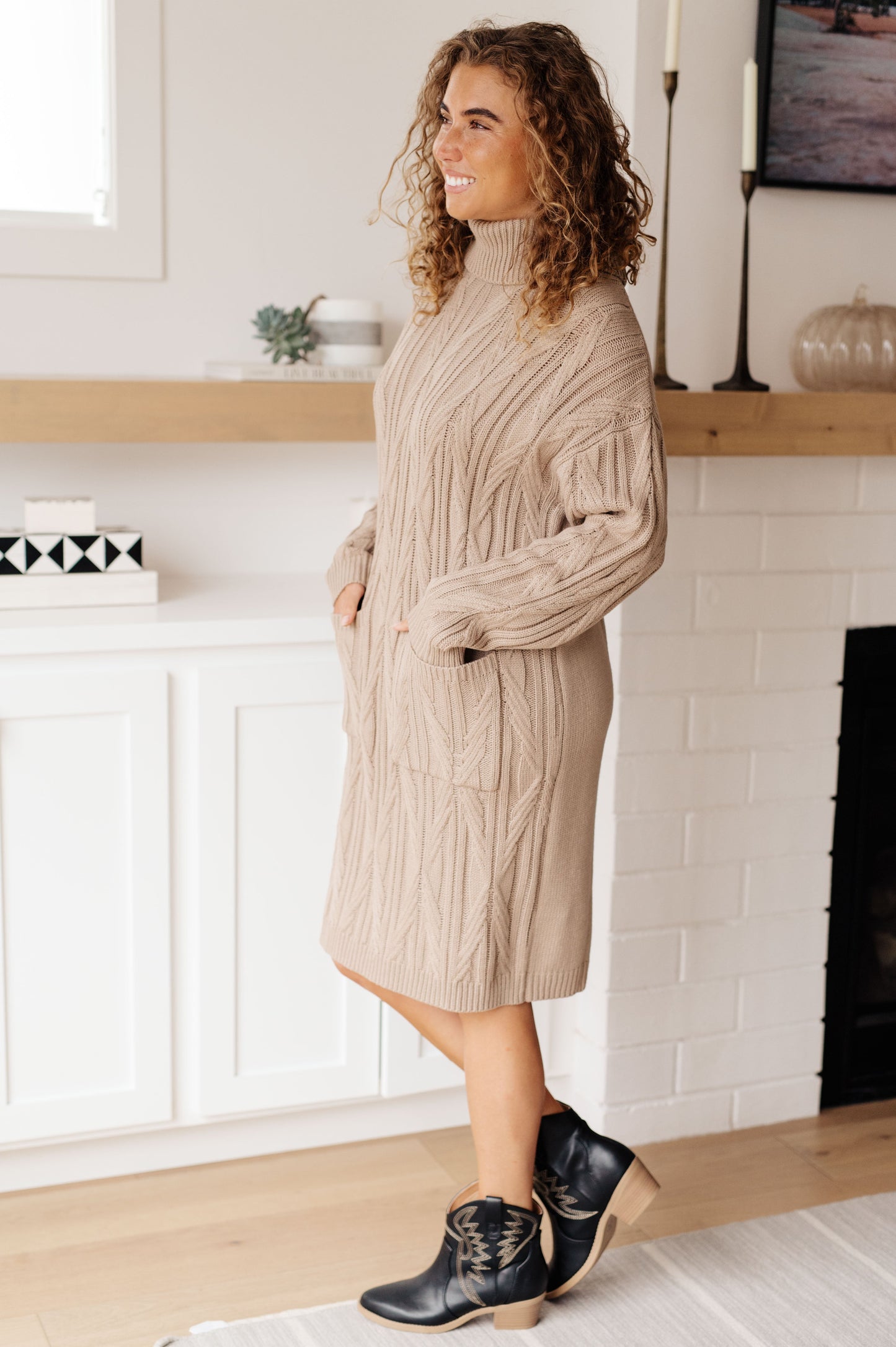Woman wearing a beige cable knit sweater dress in a home setting.