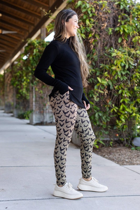 Woman wearing black long-sleeve top and patterned leggings standing on a sidewalk with greenery in the background.