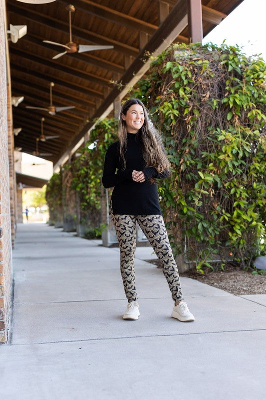 Woman in black top and patterned leggings standing on a sidewalk with greenery and a building in the background.