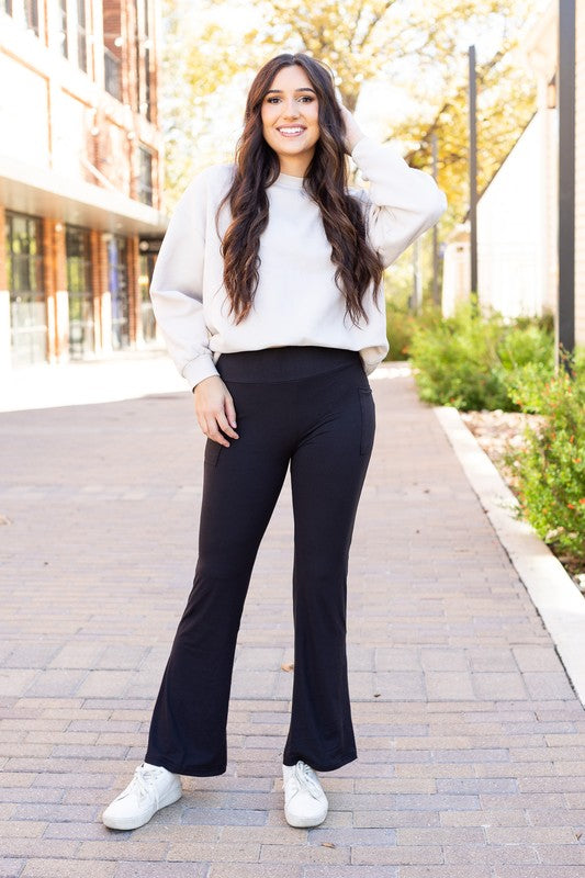 Woman wearing a white sweater and black pants standing on a sidewalk with buildings and trees in the background.