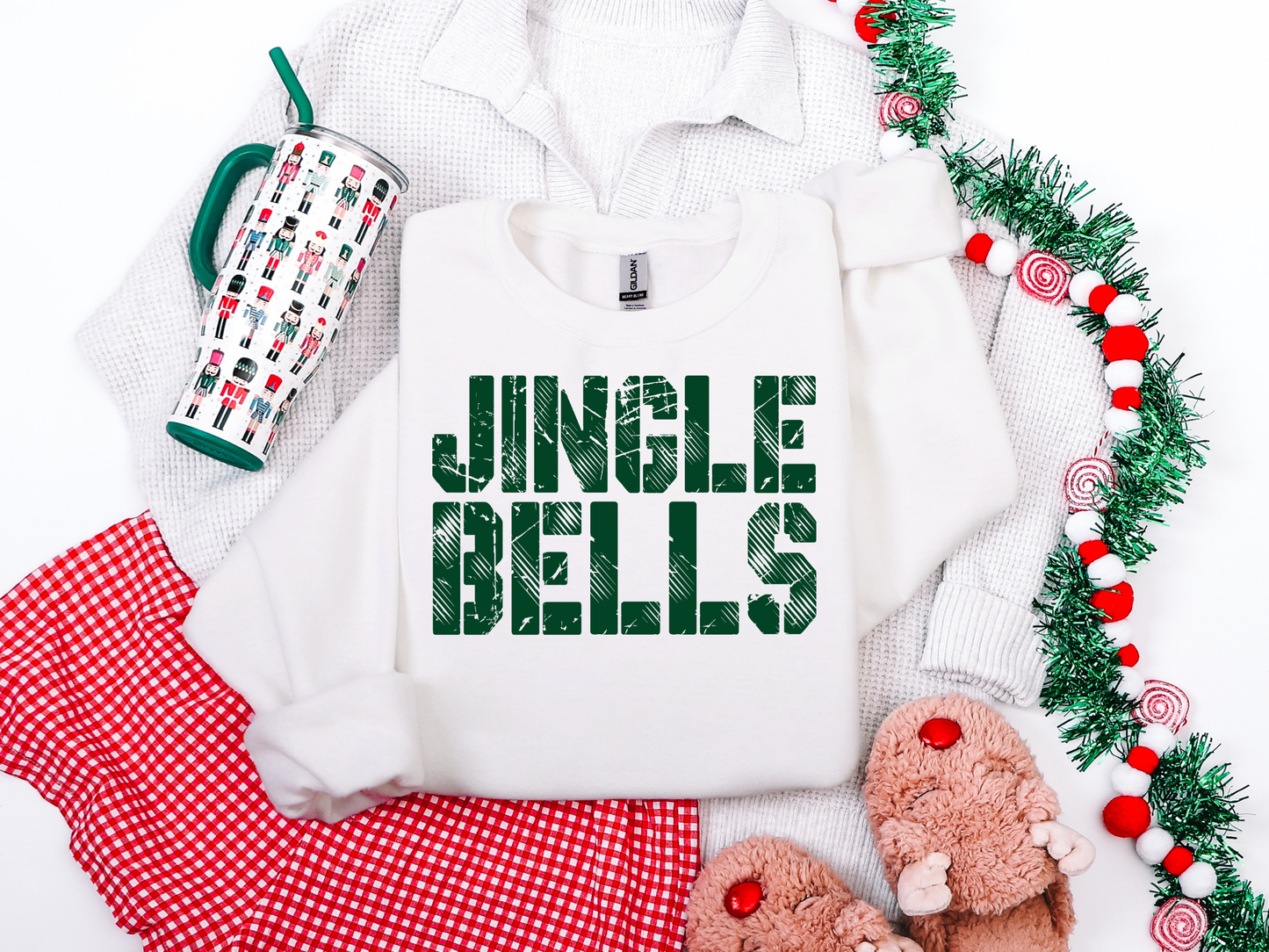 White sweatshirt with 'Jingle Bells' text, mug, red checkered blanket, and festive decor on a white background