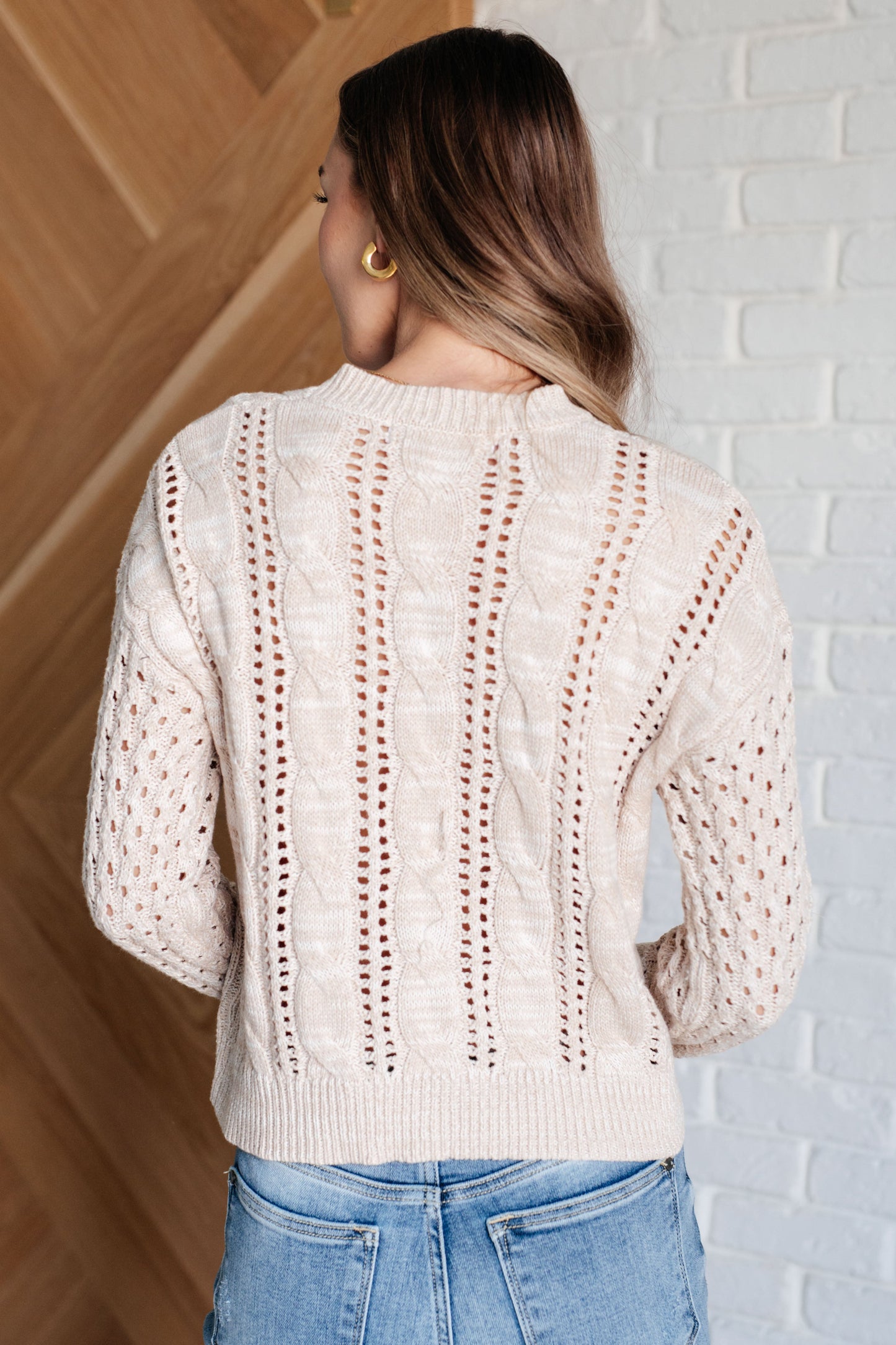 Woman wearing a cream-colored knit sweater with a textured pattern, standing against a white brick wall.