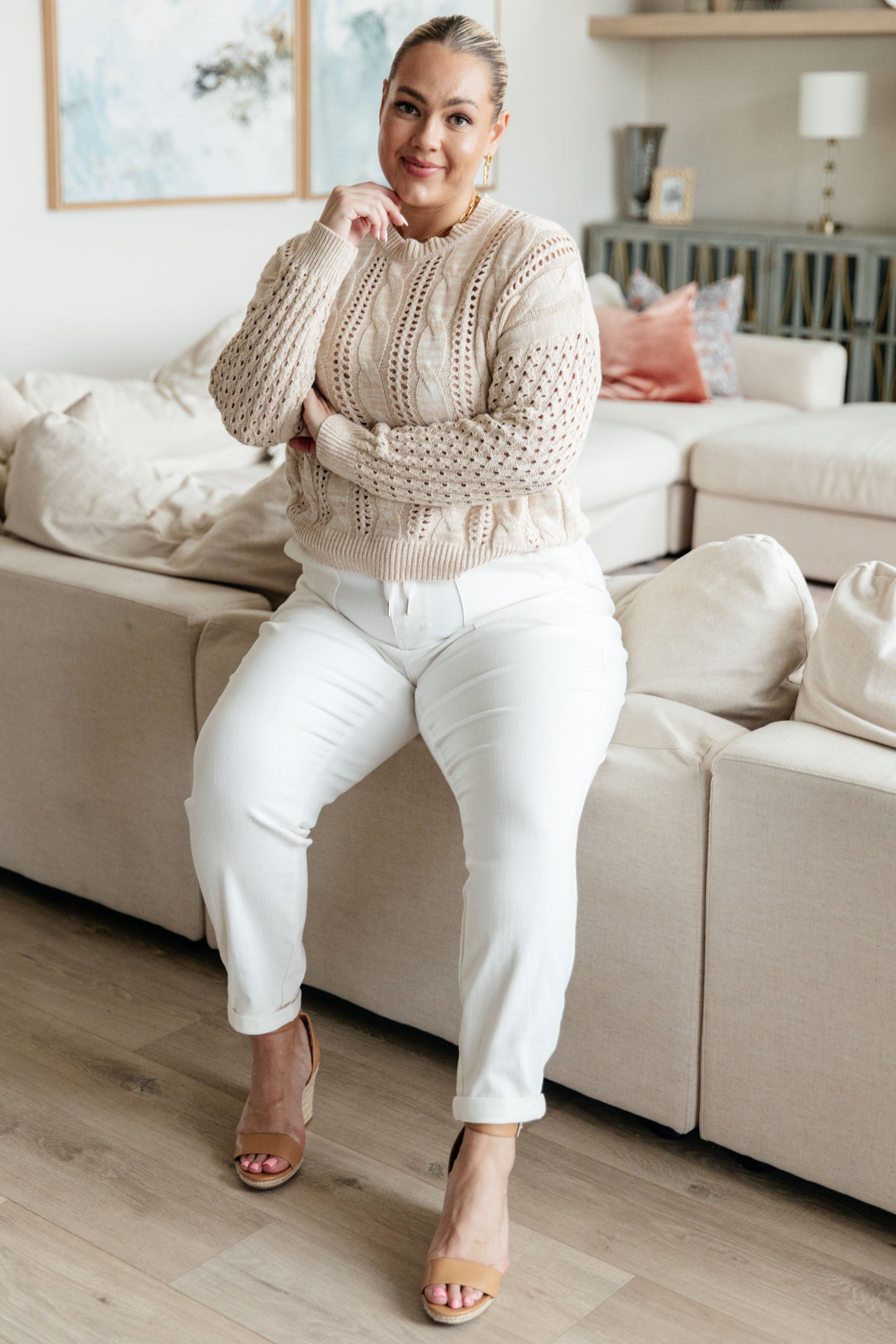 Woman sitting on a couch wearing a beige sweater and white pants in a living room.