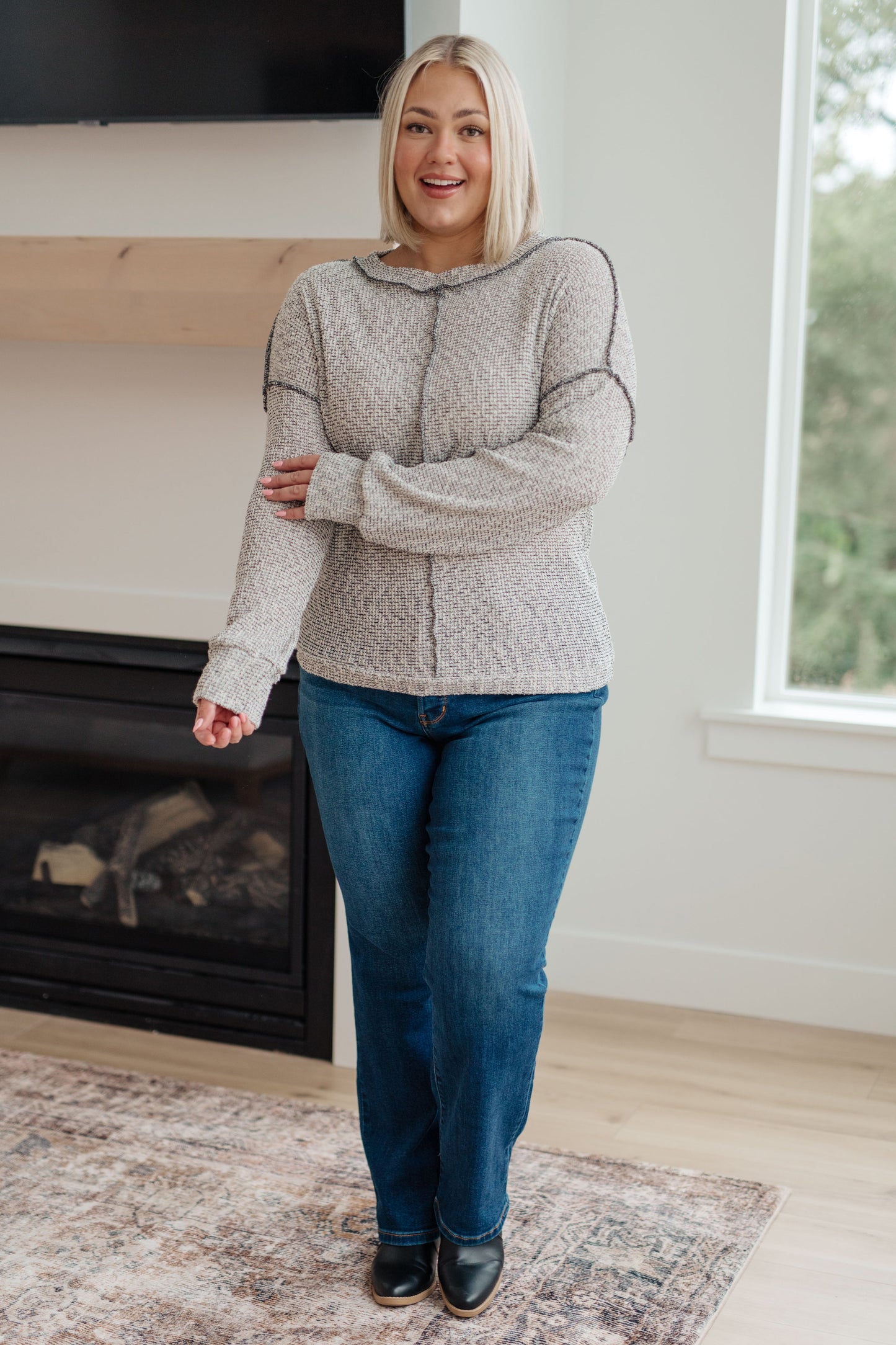 Woman wearing a patterned sweater and blue jeans standing in a room with a fireplace and window.