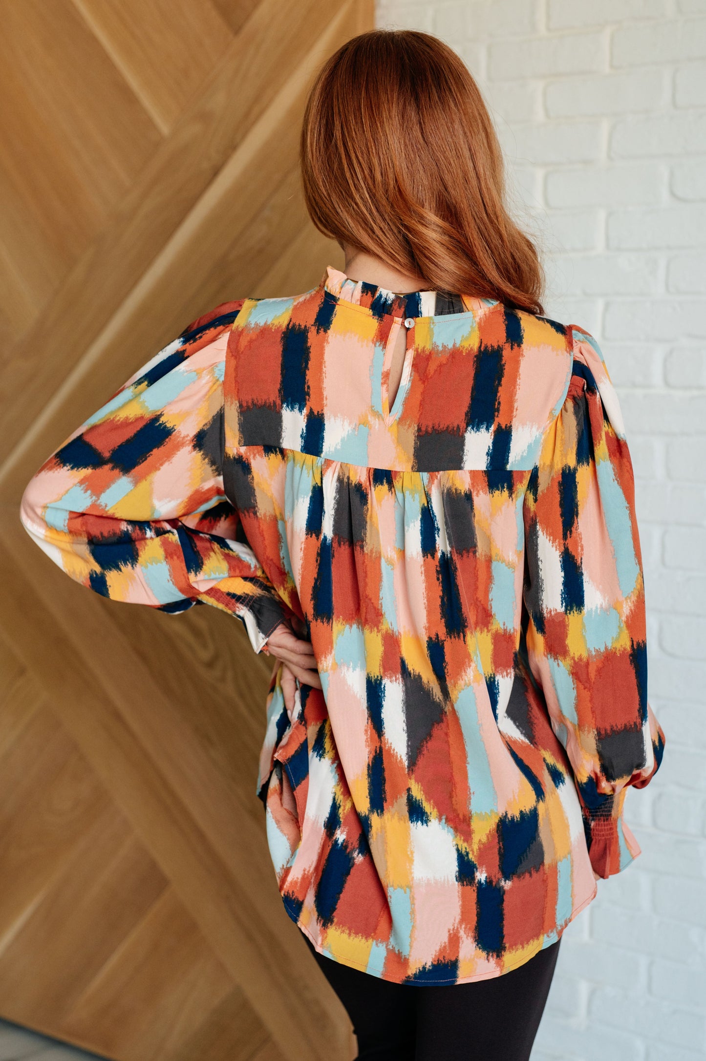 Woman wearing a colorful patterned blouse against a white brick wall.