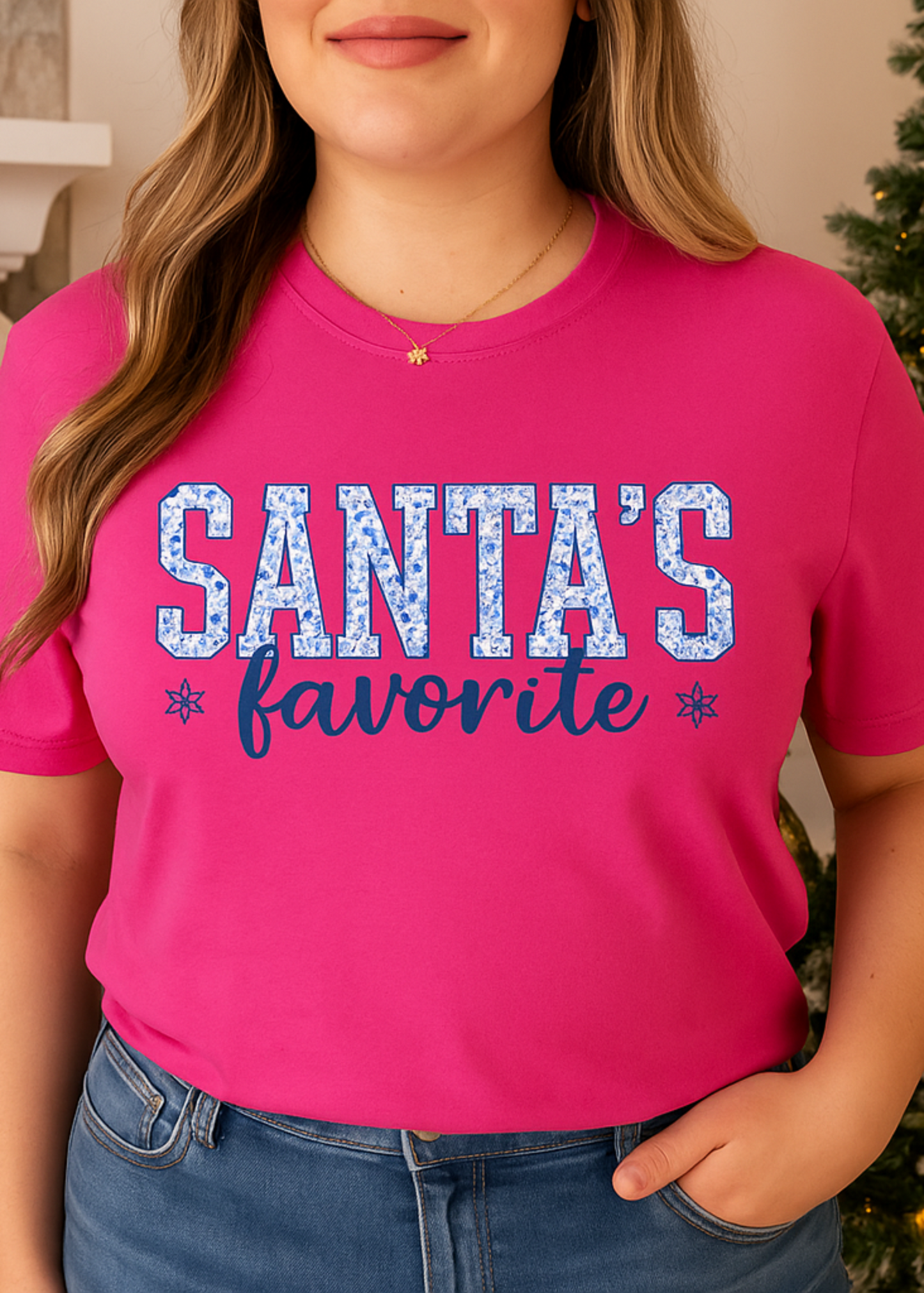 Person wearing a pink t-shirt with 'Santa's favorite' text in front of a decorated Christmas tree.