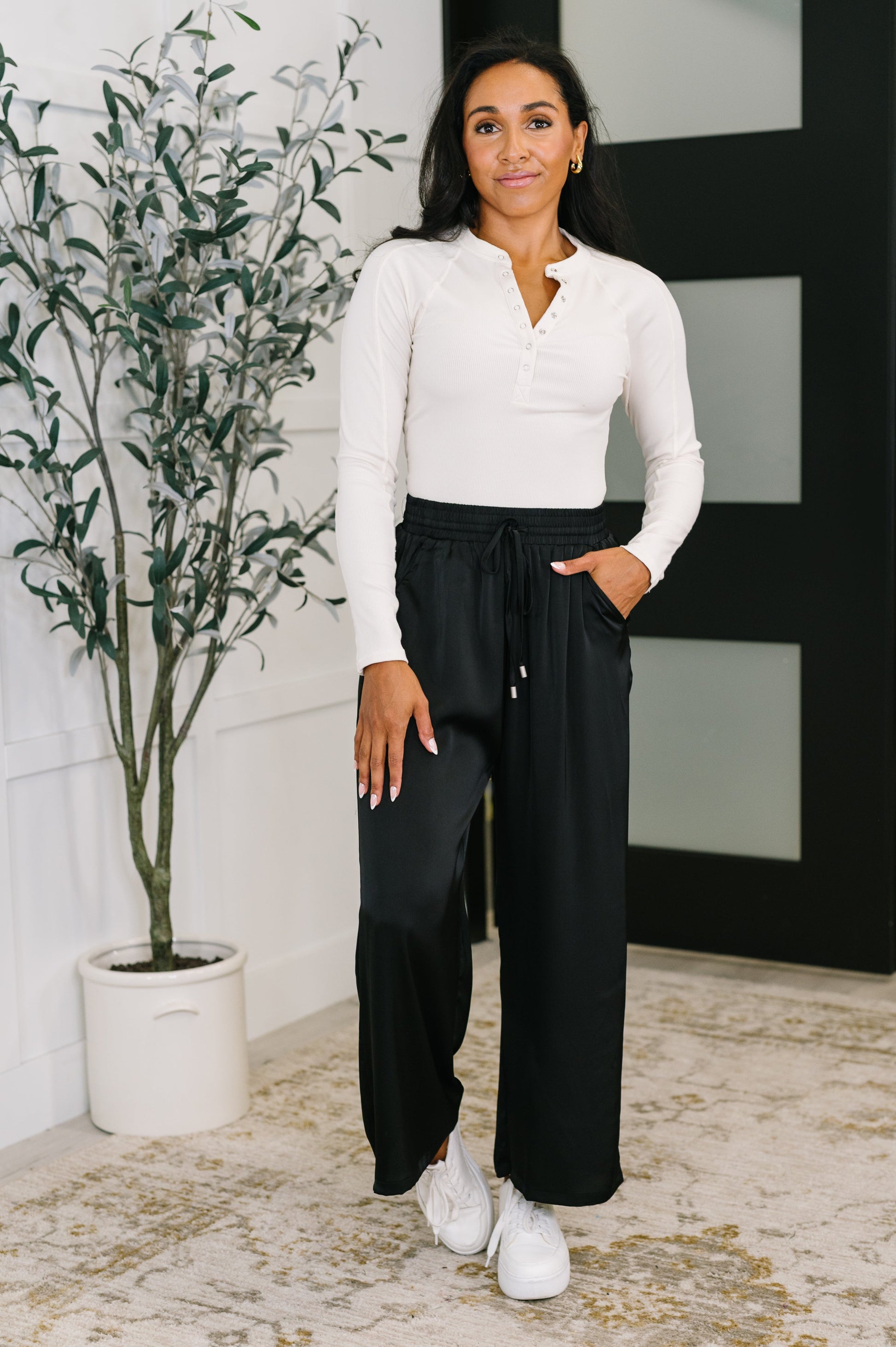 Woman wearing a white top and black pants standing indoors with a plant in the background