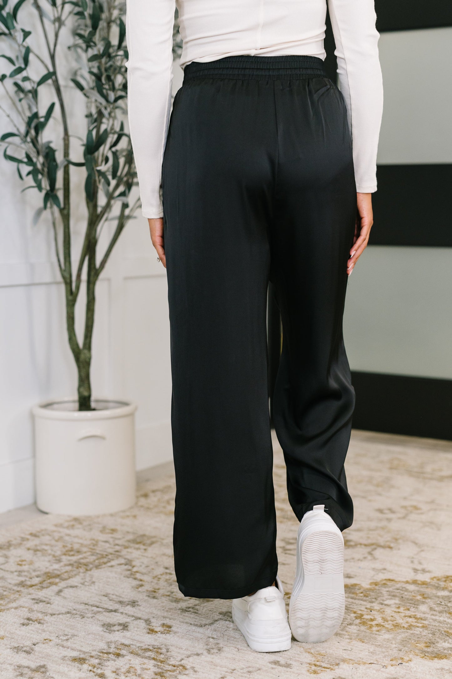Person wearing black pants and white sneakers in a room with a plant and rug.