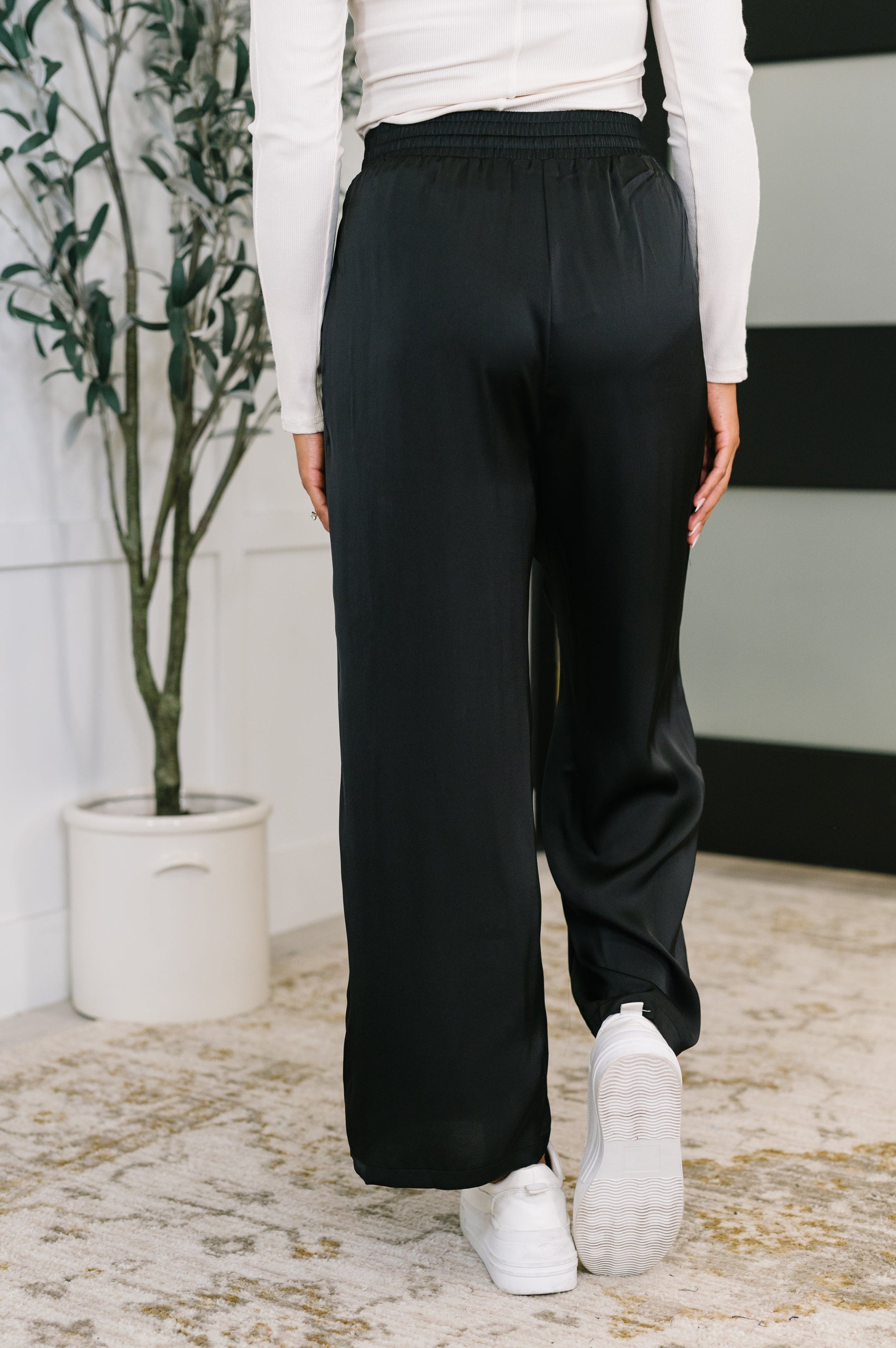 Person wearing black pants and white sneakers in a room with a plant and rug.