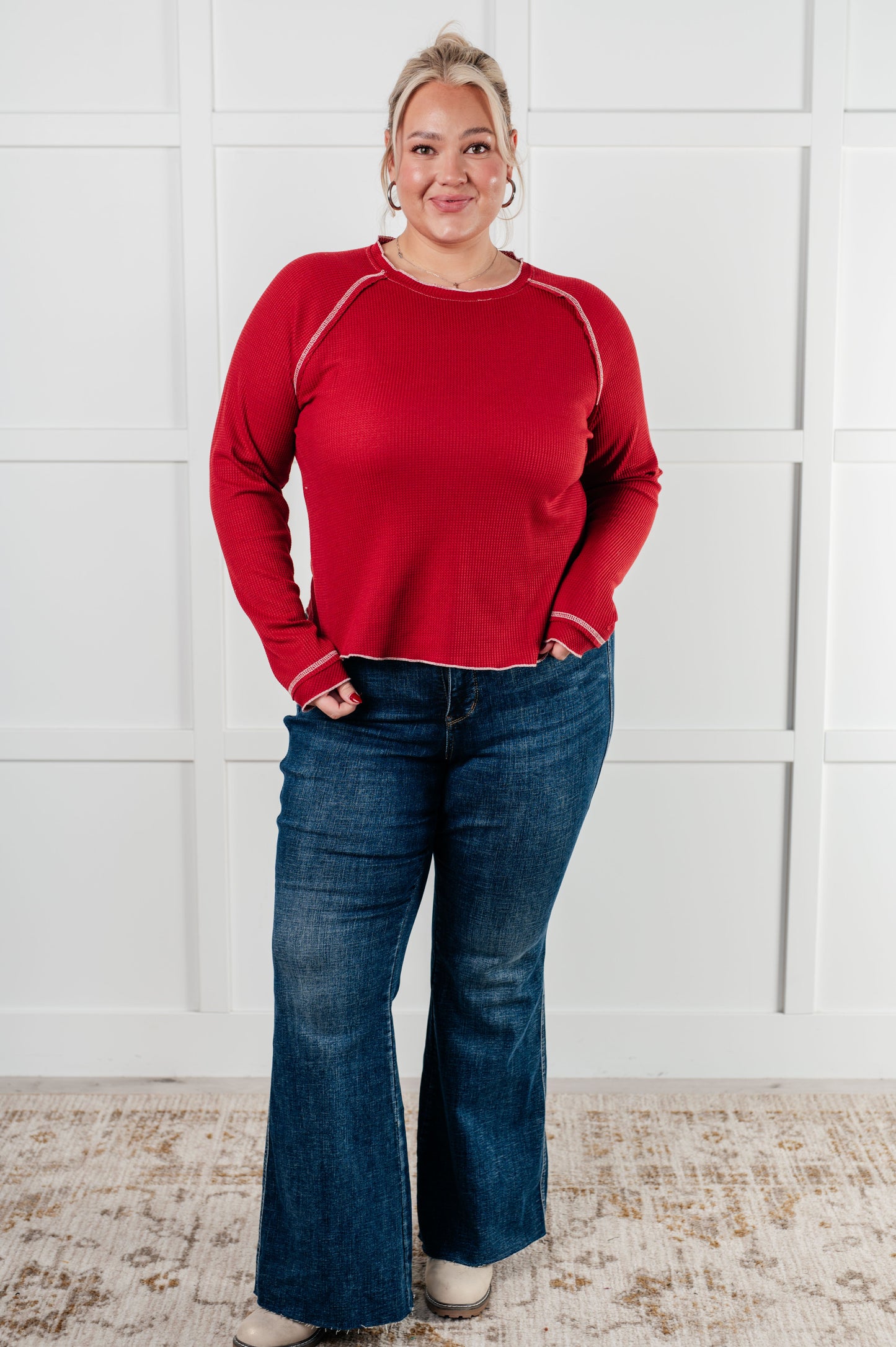 Person wearing a red sweater and blue jeans standing against a white paneled wall.