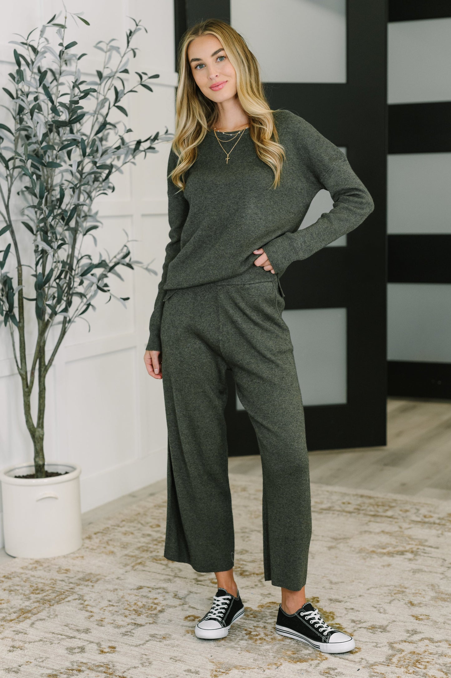 Woman wearing a green outfit standing indoors with a plant and black and white striped wall in the background.