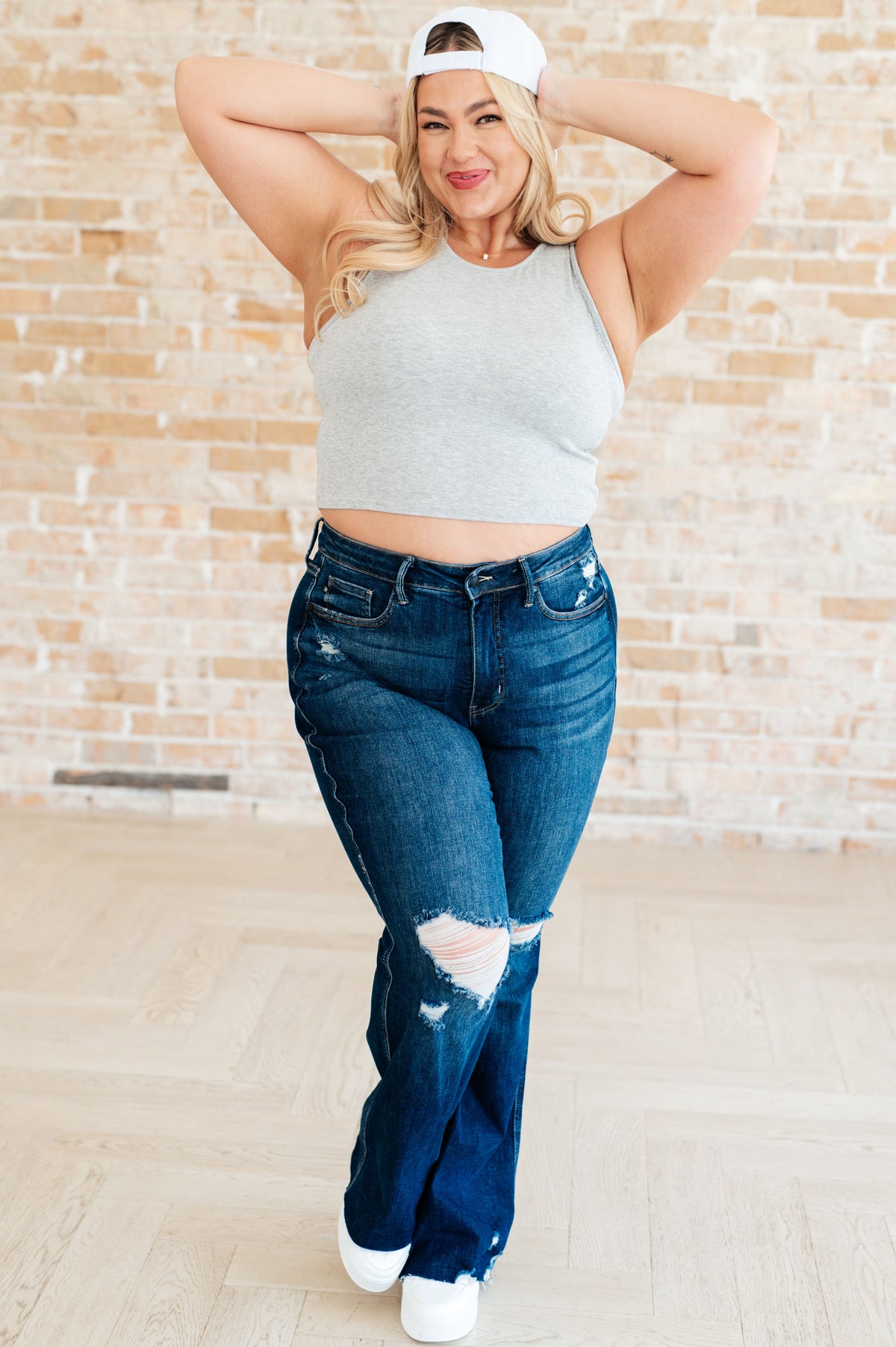 Woman wearing a gray crop top and blue jeans with a white cap, standing against a beige brick wall.