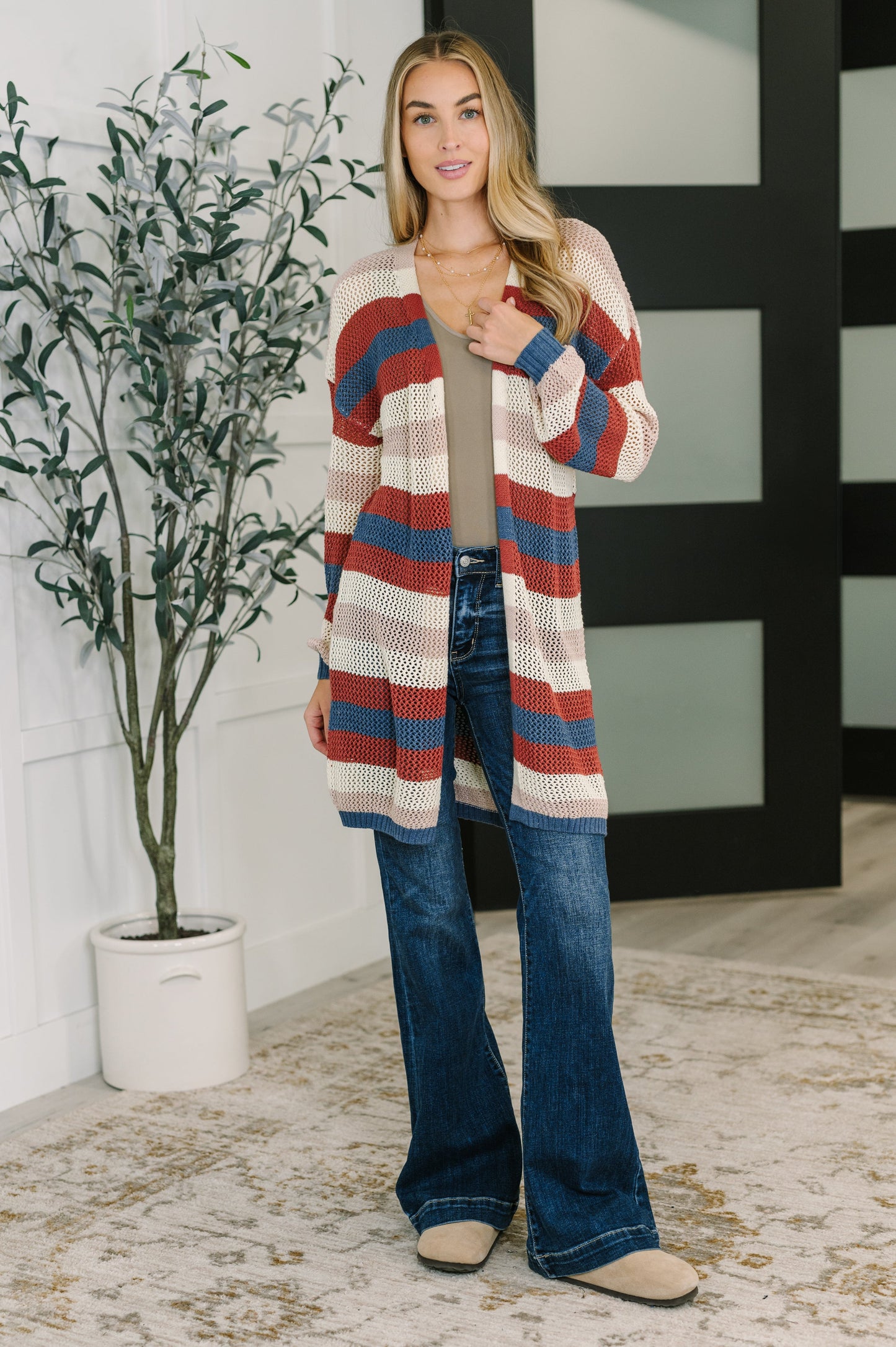 Woman wearing a striped cardigan and jeans standing indoors next to a plant.