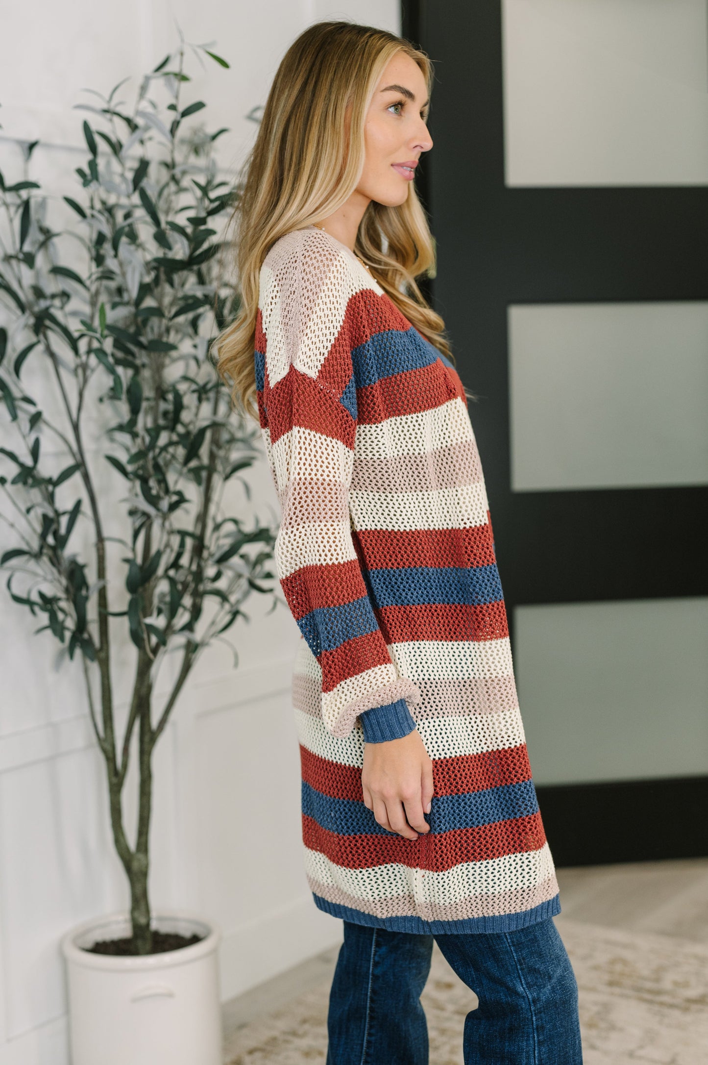 Woman wearing a striped cardigan standing indoors next to a plant.