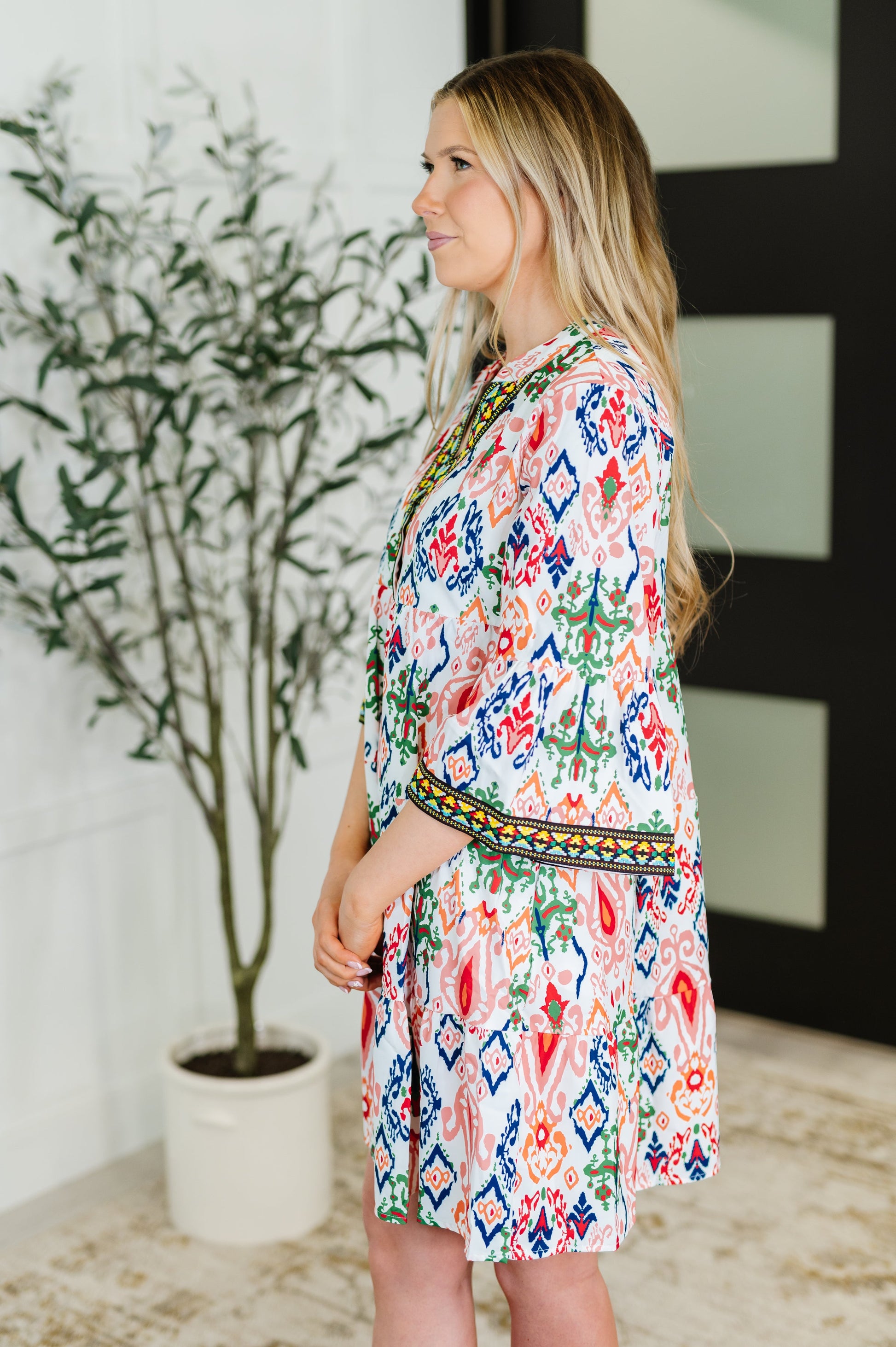 Woman wearing a colorful patterned dress standing indoors with a plant in the background