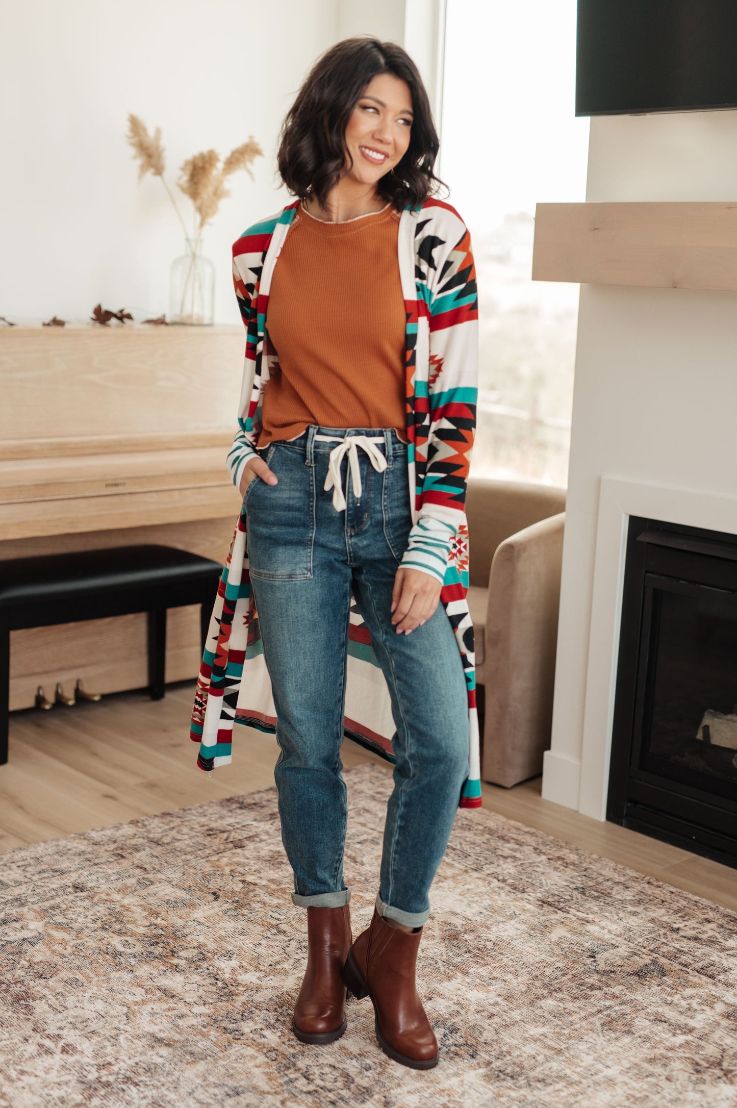 Woman wearing a colorful cardigan, brown shirt, blue jeans, and brown boots in a living room.