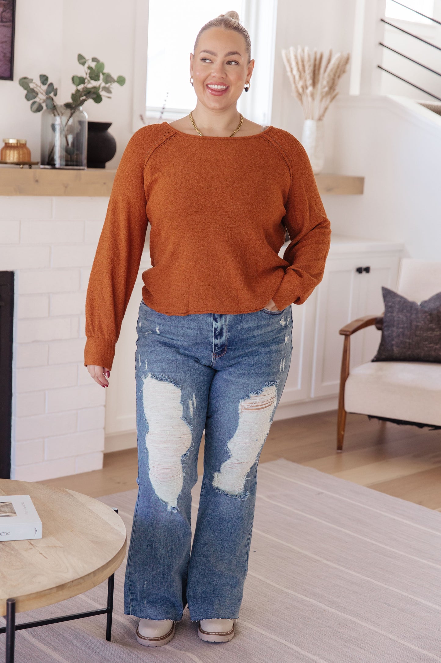 Woman wearing a rust-colored sweater and blue jeans in a living room setting.