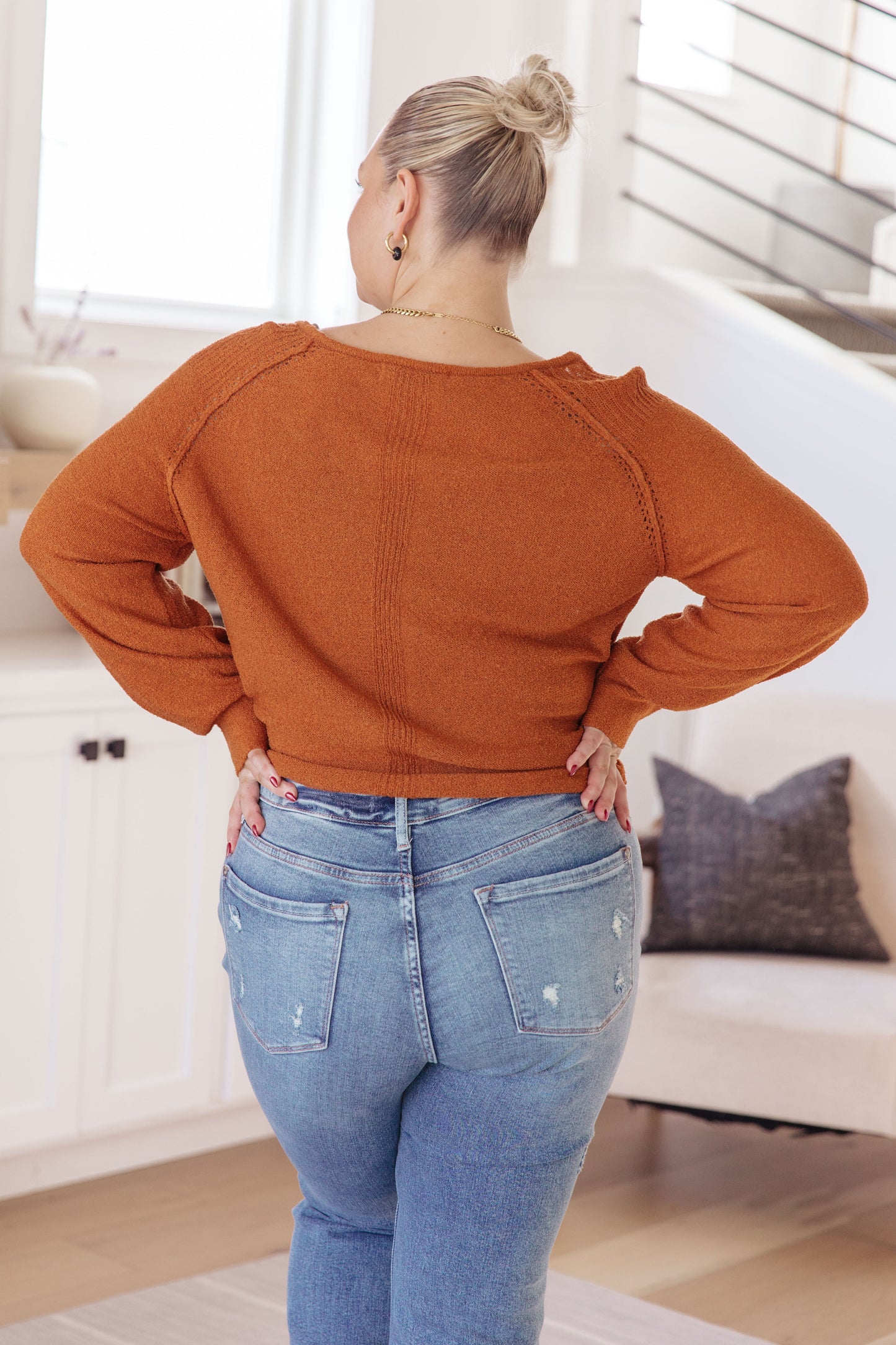 Person wearing a rust-colored sweater and blue jeans in a room with a couch and pillow.