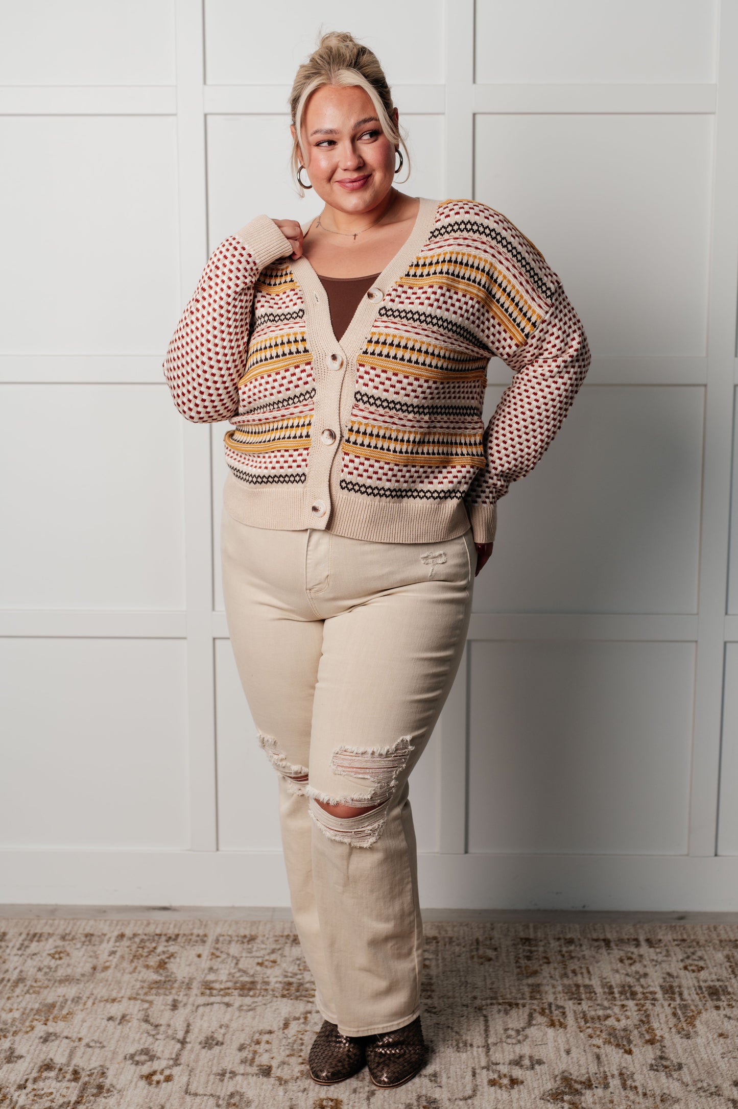 Woman wearing a patterned cardigan over a beige top and pants, standing against a white paneled wall.