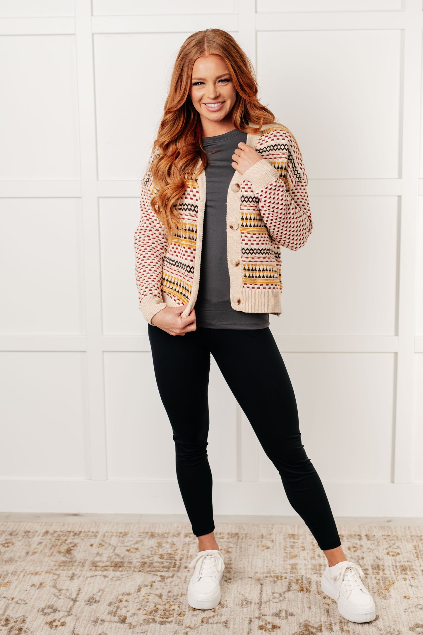 Woman wearing a patterned cardigan, gray shirt, black leggings, and white sneakers against a white wall.