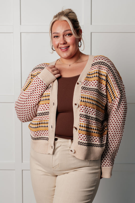 Woman wearing a patterned cardigan over a brown top and beige pants against a white tiled wall.