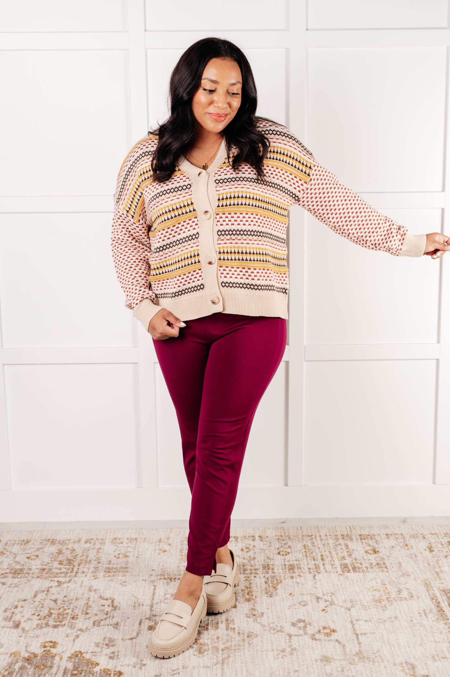 Woman wearing a patterned sweater and red pants against a white wall.