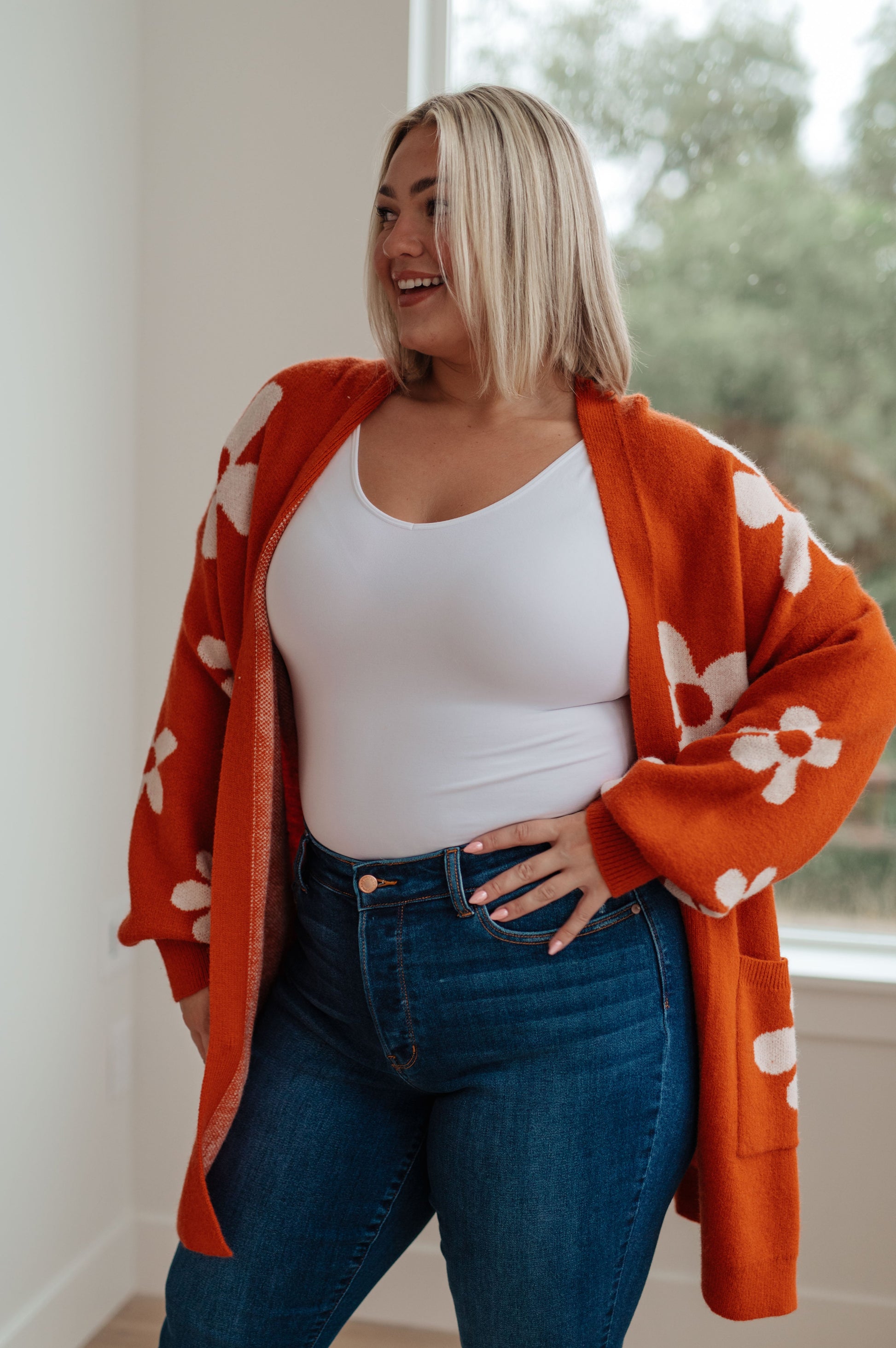 Woman wearing a white top and blue jeans with an orange floral cardigan indoors.