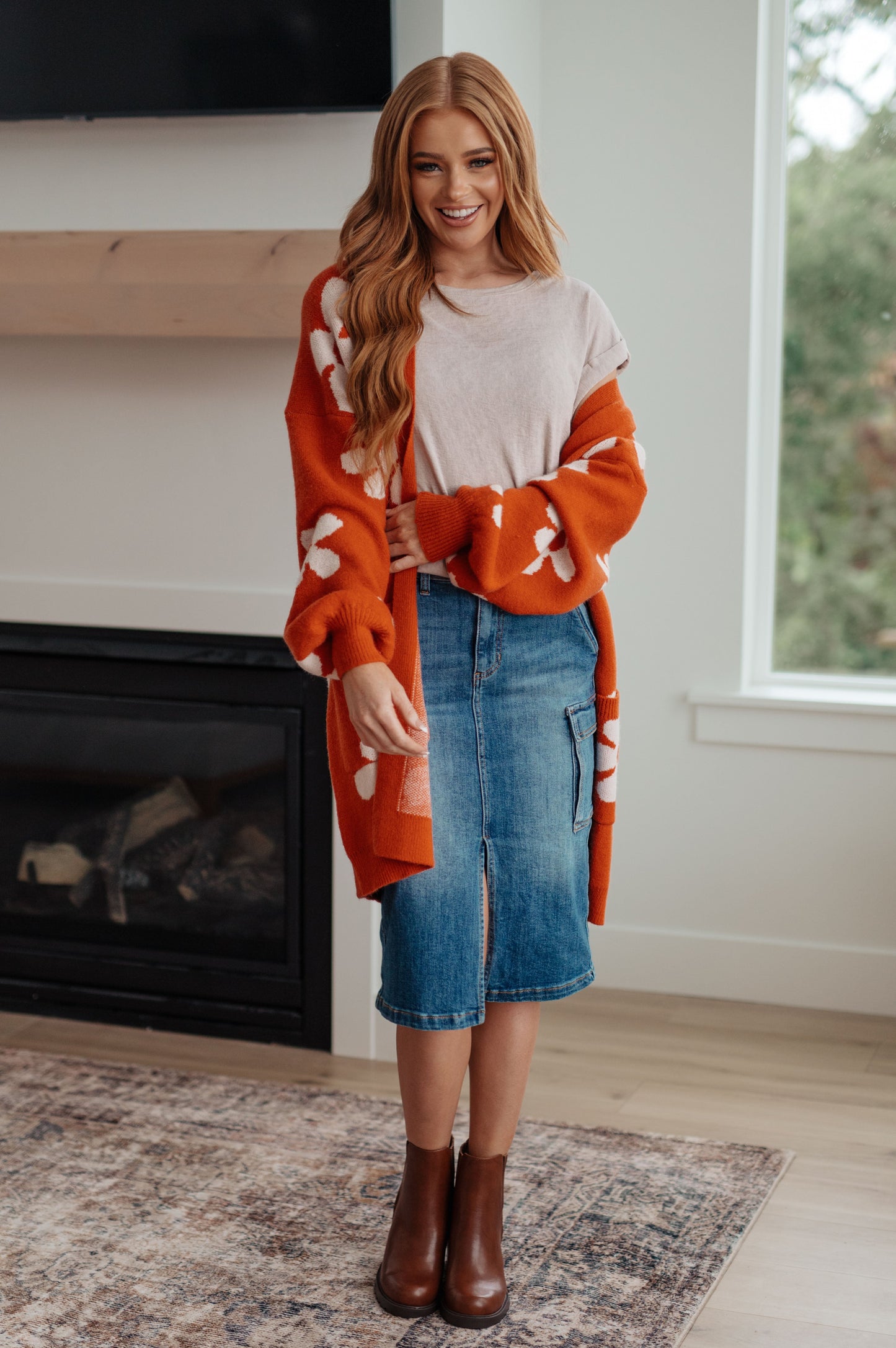Woman wearing an orange cardigan with white patterns, beige top, blue skirt, and brown boots in a room with a fireplace.