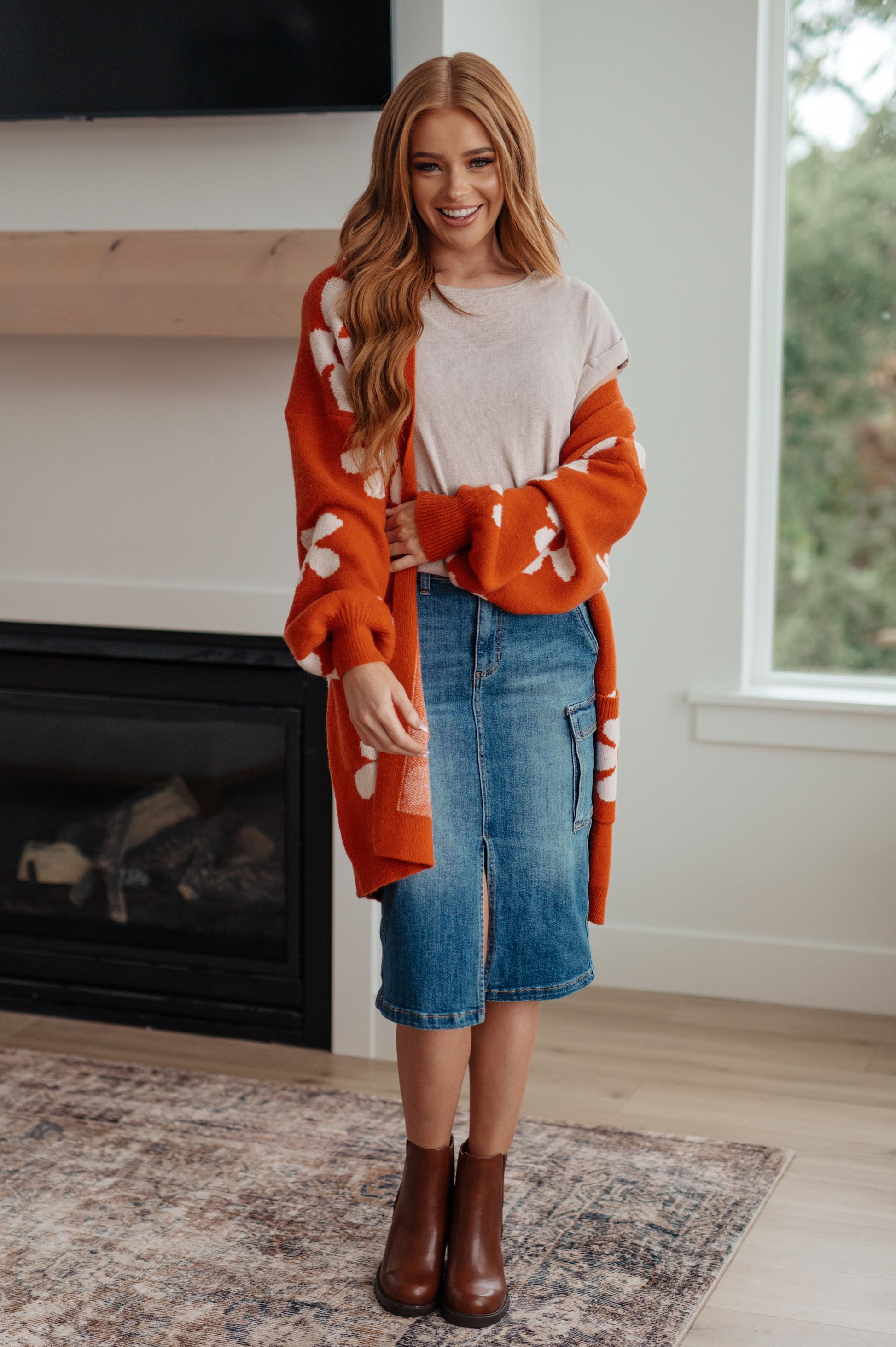 Woman wearing an orange cardigan with white patterns, beige top, blue skirt, and brown boots in a room with a fireplace.