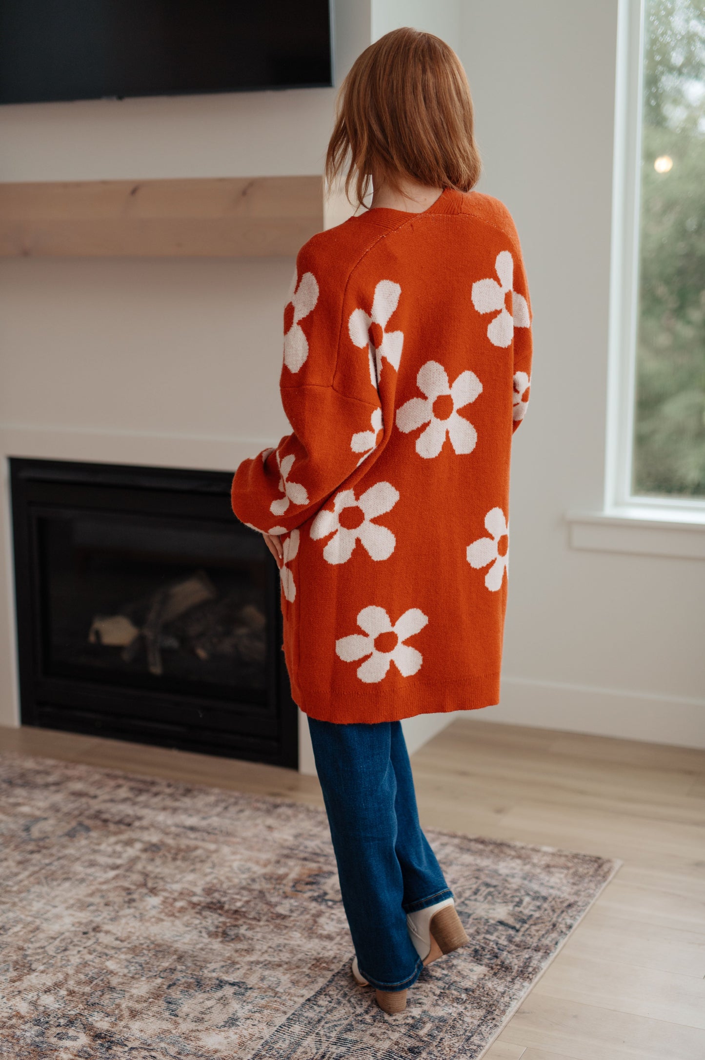 Person wearing a red sweater with white floral patterns in a room with a fireplace and window.