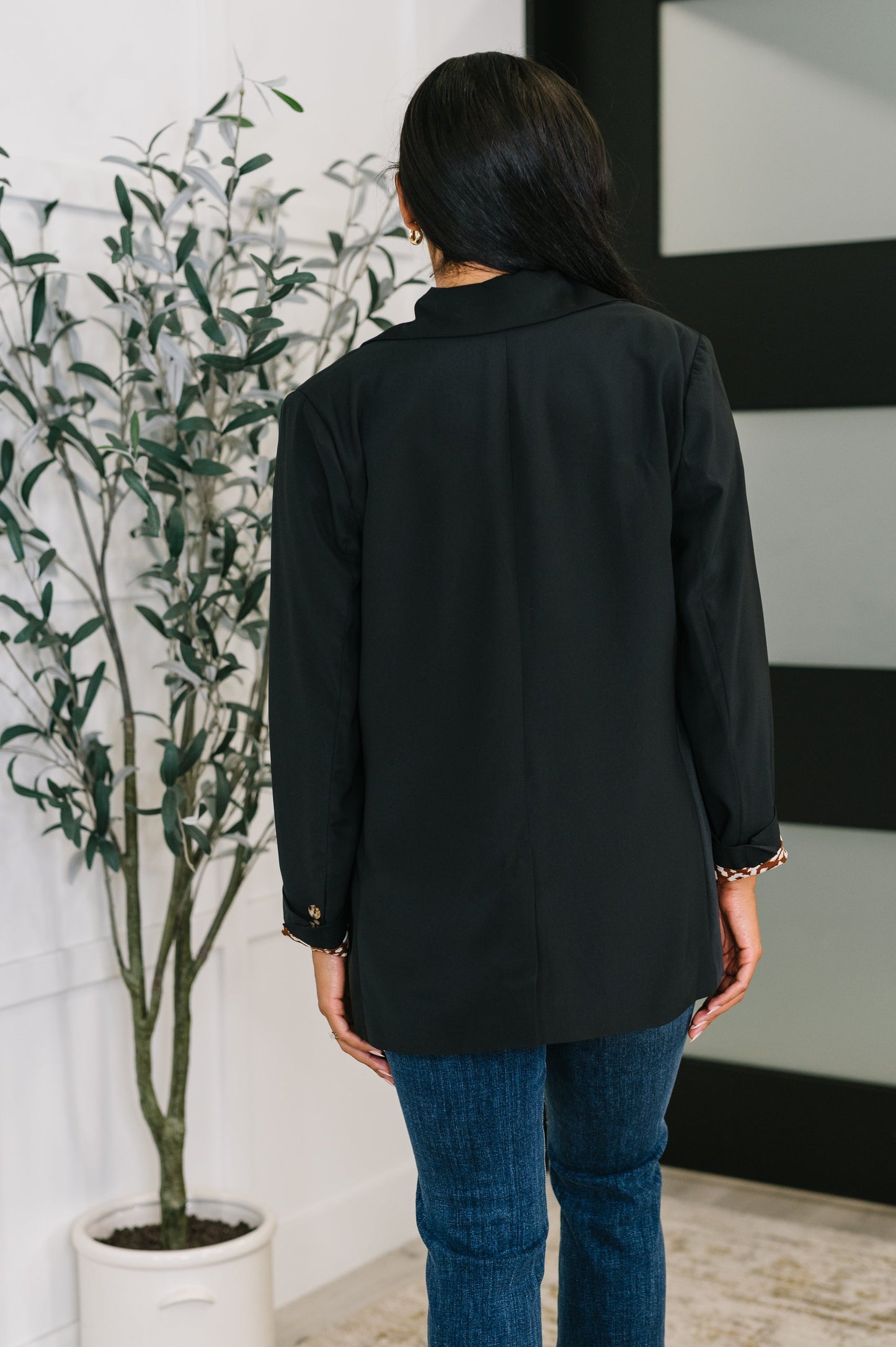 Person wearing a black shirt and blue jeans standing in a room with a plant and wall art.