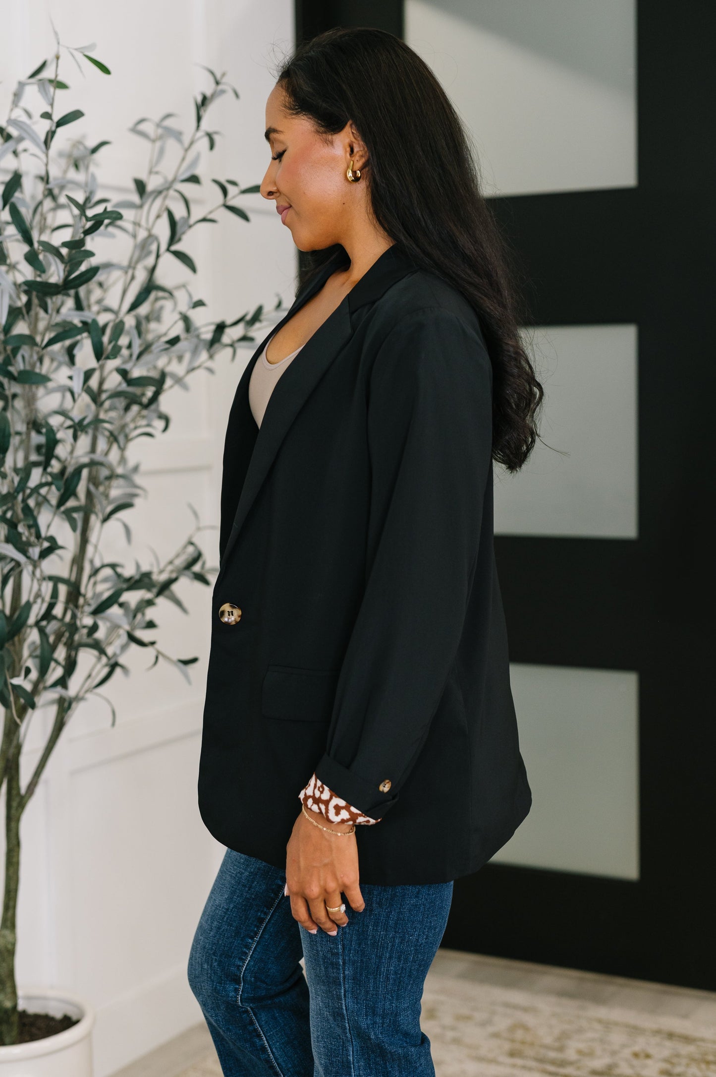 Woman wearing a black blazer and blue jeans standing indoors with a plant and abstract art in the background.