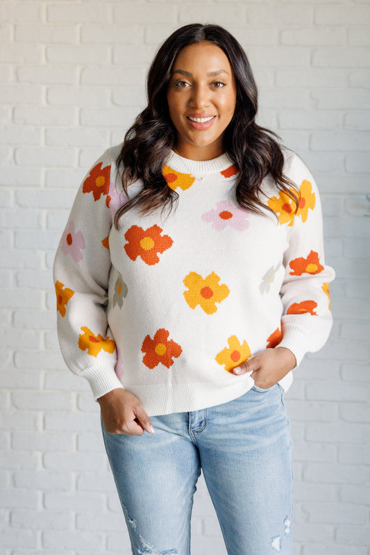 Woman wearing a white sweater with orange floral patterns against a white brick wall.