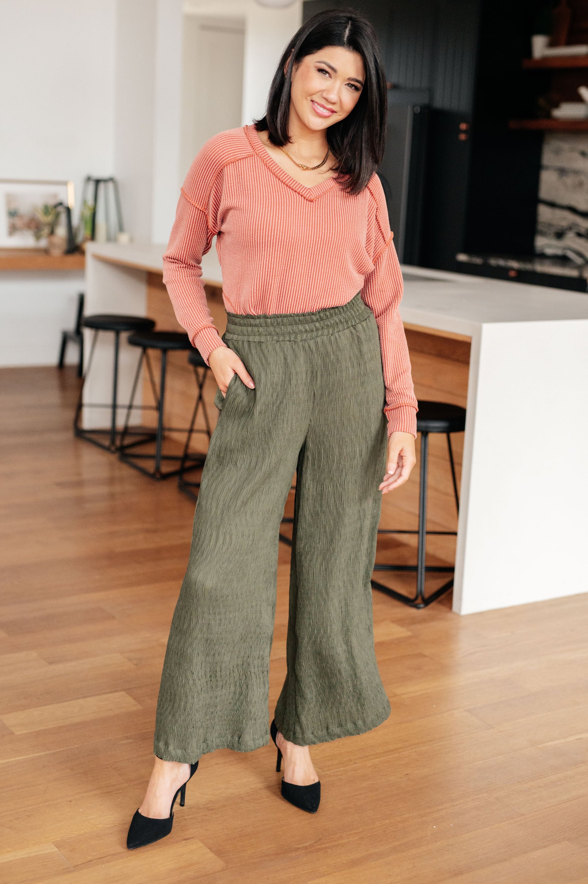 Woman wearing a pink sweater and green pants in a kitchen.