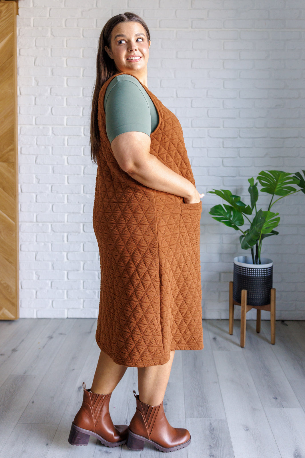 Woman wearing a brown quilted dress and boots in a room with a white brick wall and plant.