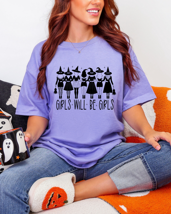 Person wearing a purple t-shirt with 'Girls Will Be Girls' text and black witch graphics, sitting on an orange pillow.