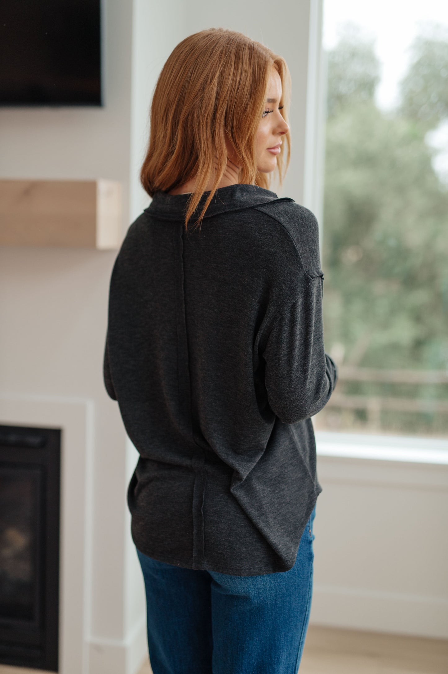 Woman wearing a dark gray hoodie and blue jeans standing in a room with a fireplace and window.
