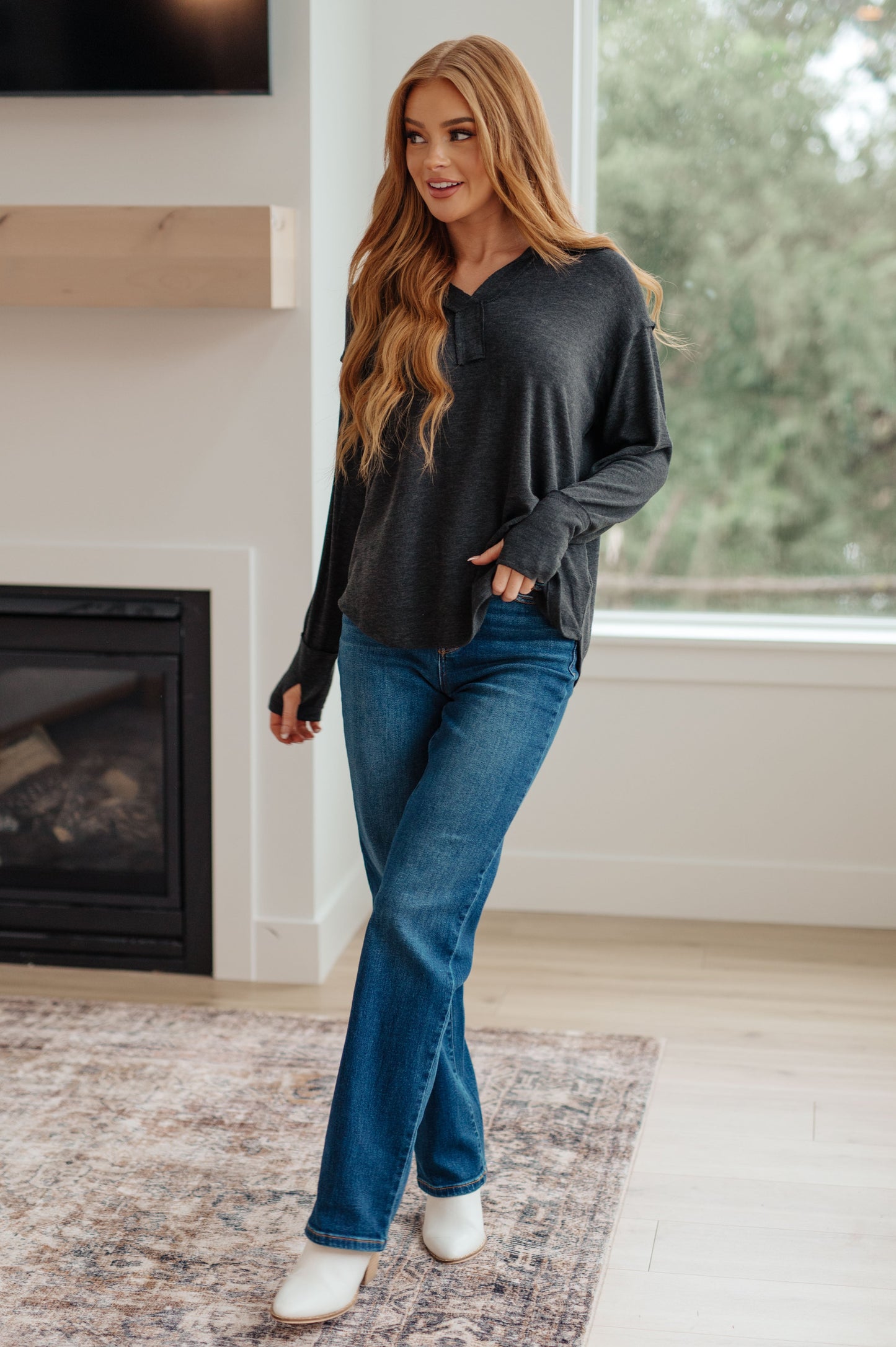 Woman wearing a black long-sleeve top and blue jeans standing in a room with a fireplace and large window.
