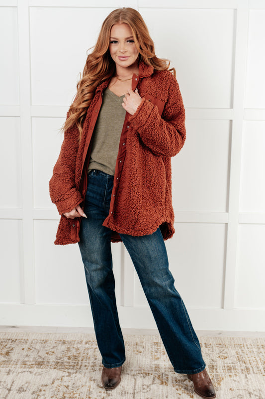 Woman wearing a rust-colored coat, green shirt, and blue jeans against a white wall.