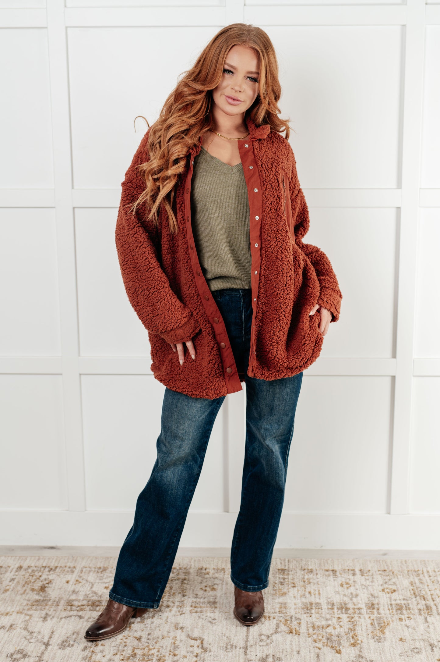 Woman wearing a rust-colored cardigan over a green shirt and blue jeans against a white wall.