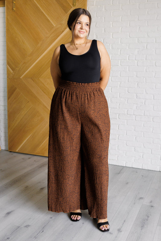 Woman wearing a black top and brown wide-leg pants standing against a white brick wall.