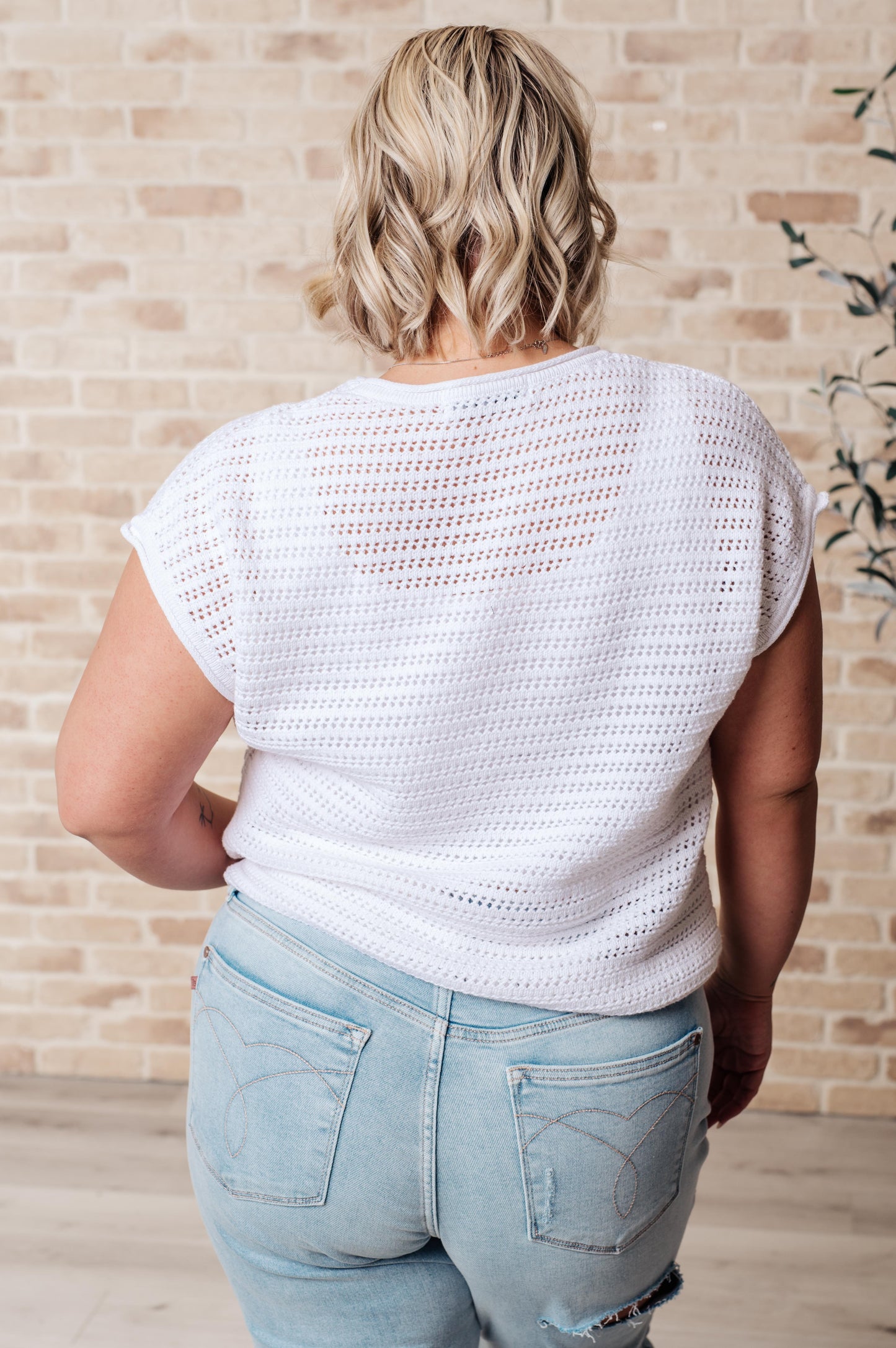 Person wearing a white crochet top and light blue jeans against a beige brick wall.