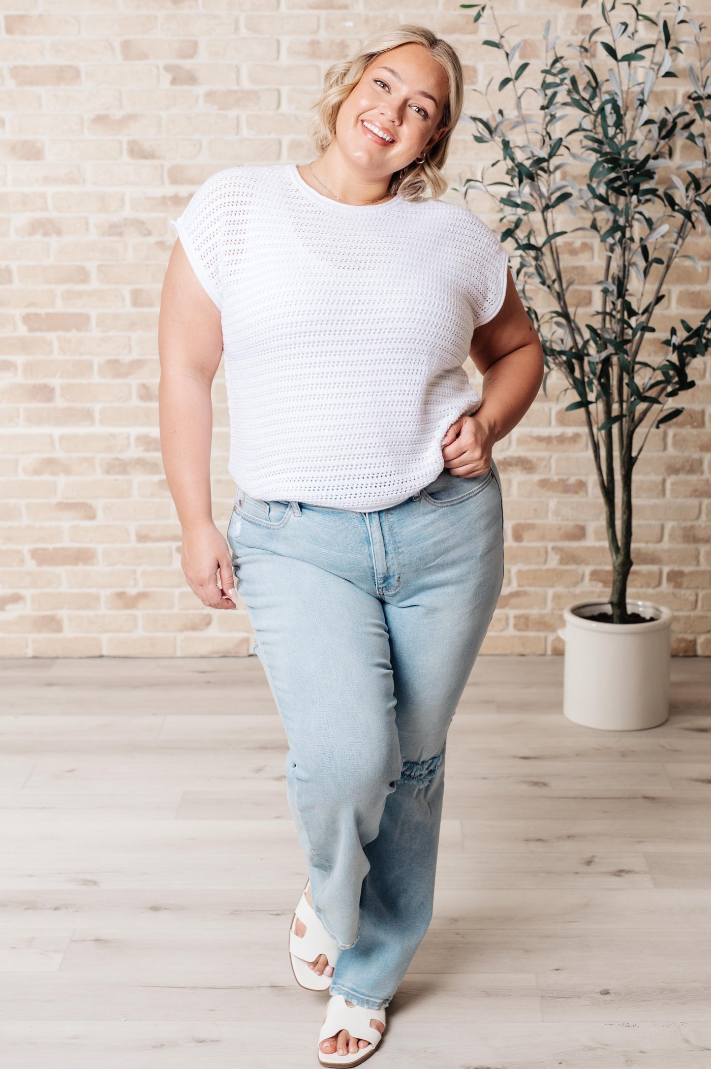 Woman wearing a white knit top and light blue jeans standing in a room with a plant and brick wall.