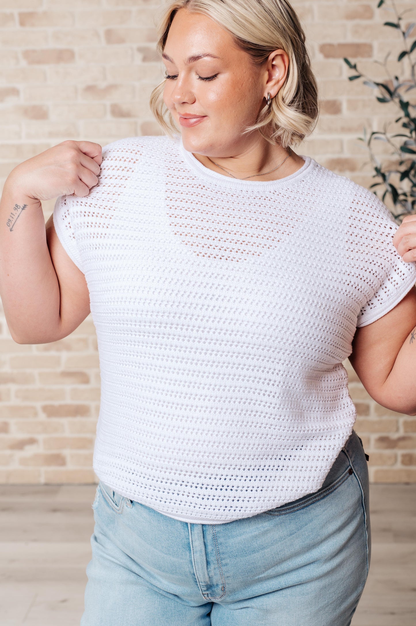 A woman wearing a white open knit cap sleeve sweater with a round neckline, paired with blue jeans.