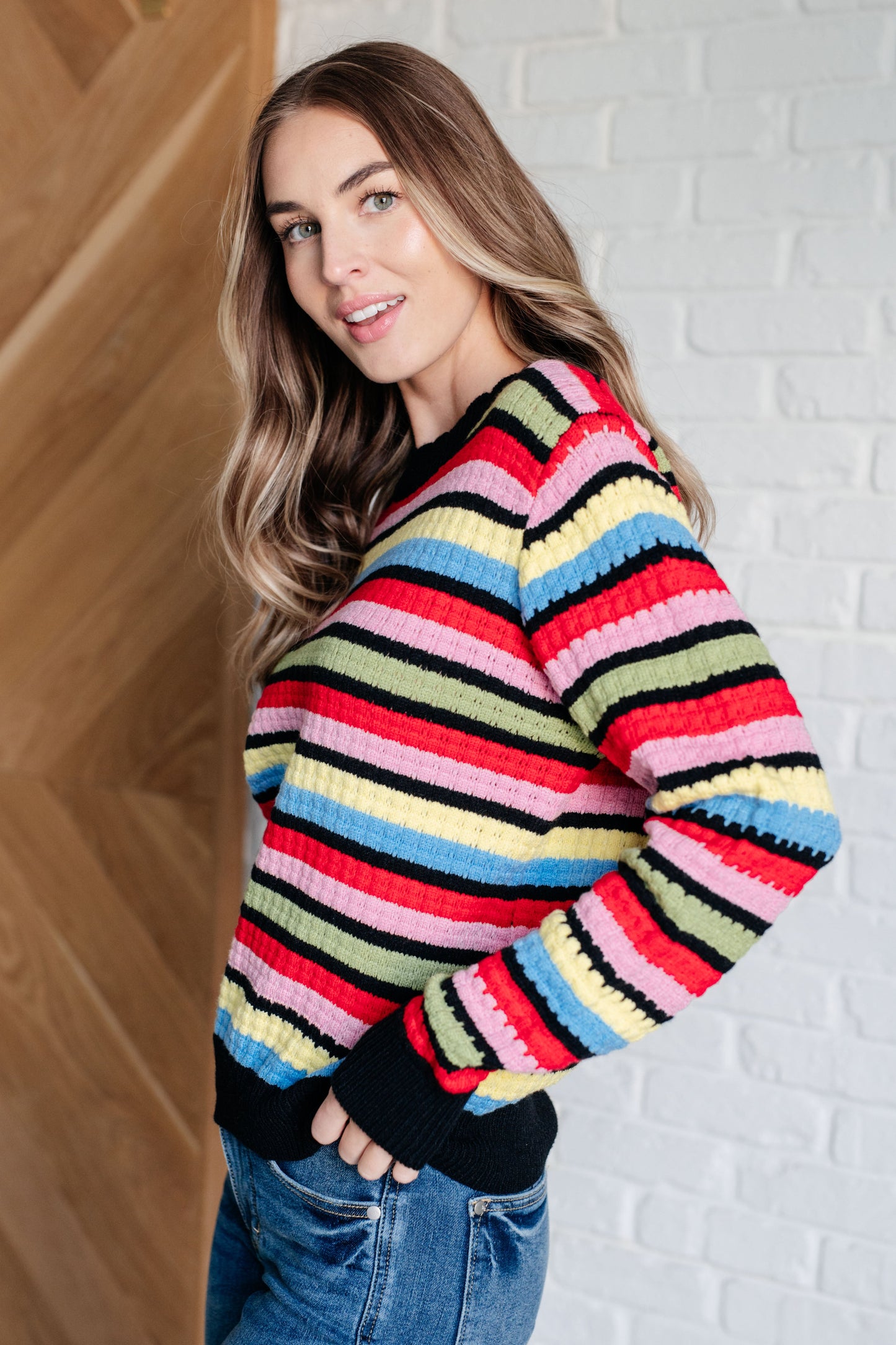 Woman wearing a colorful striped sweater against a white brick wall.
