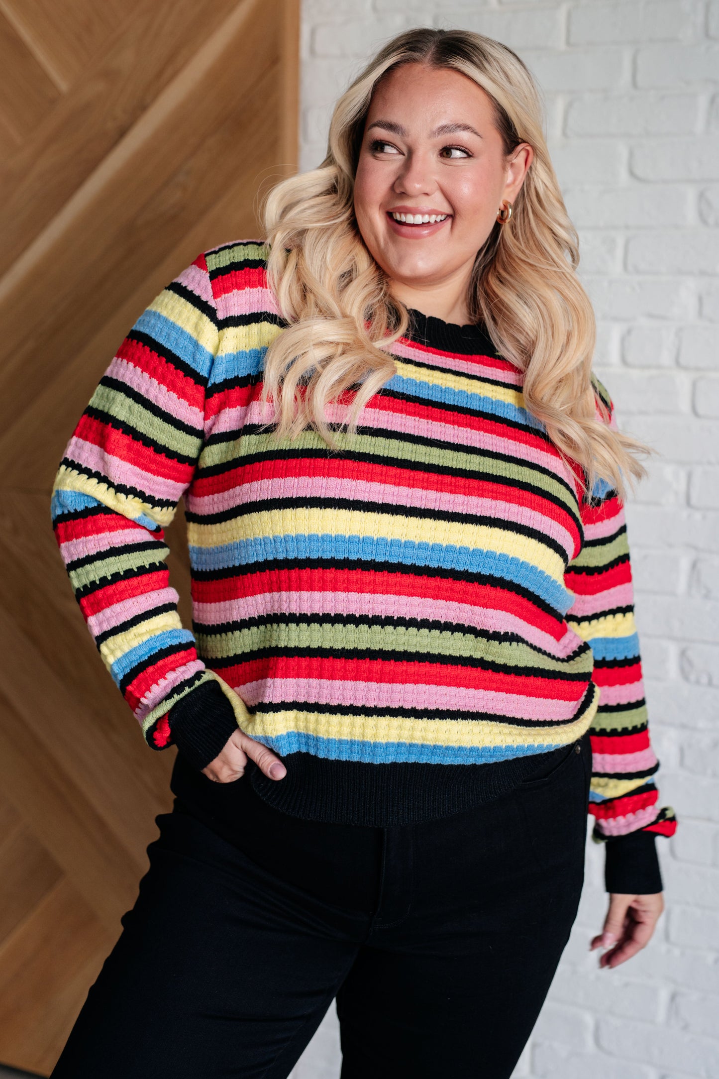 Woman wearing a colorful striped sweater against a white brick wall.