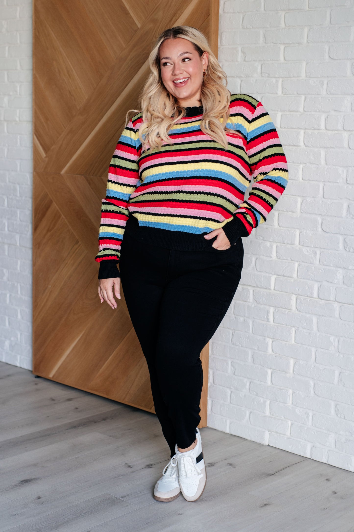 Woman wearing a colorful striped sweater and black pants standing against a white brick wall.