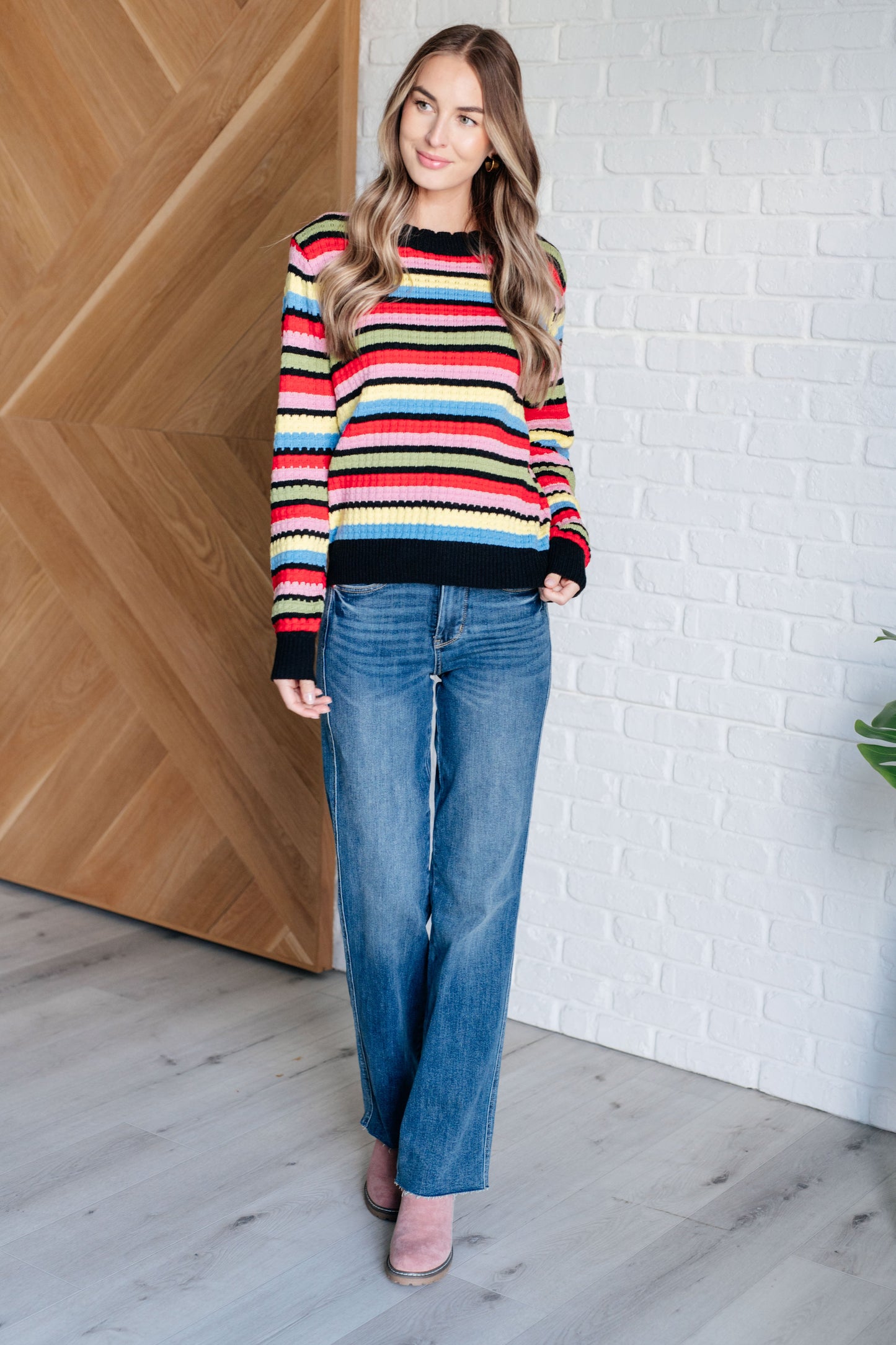 Woman wearing a colorful striped sweater and blue jeans standing against a white brick wall.