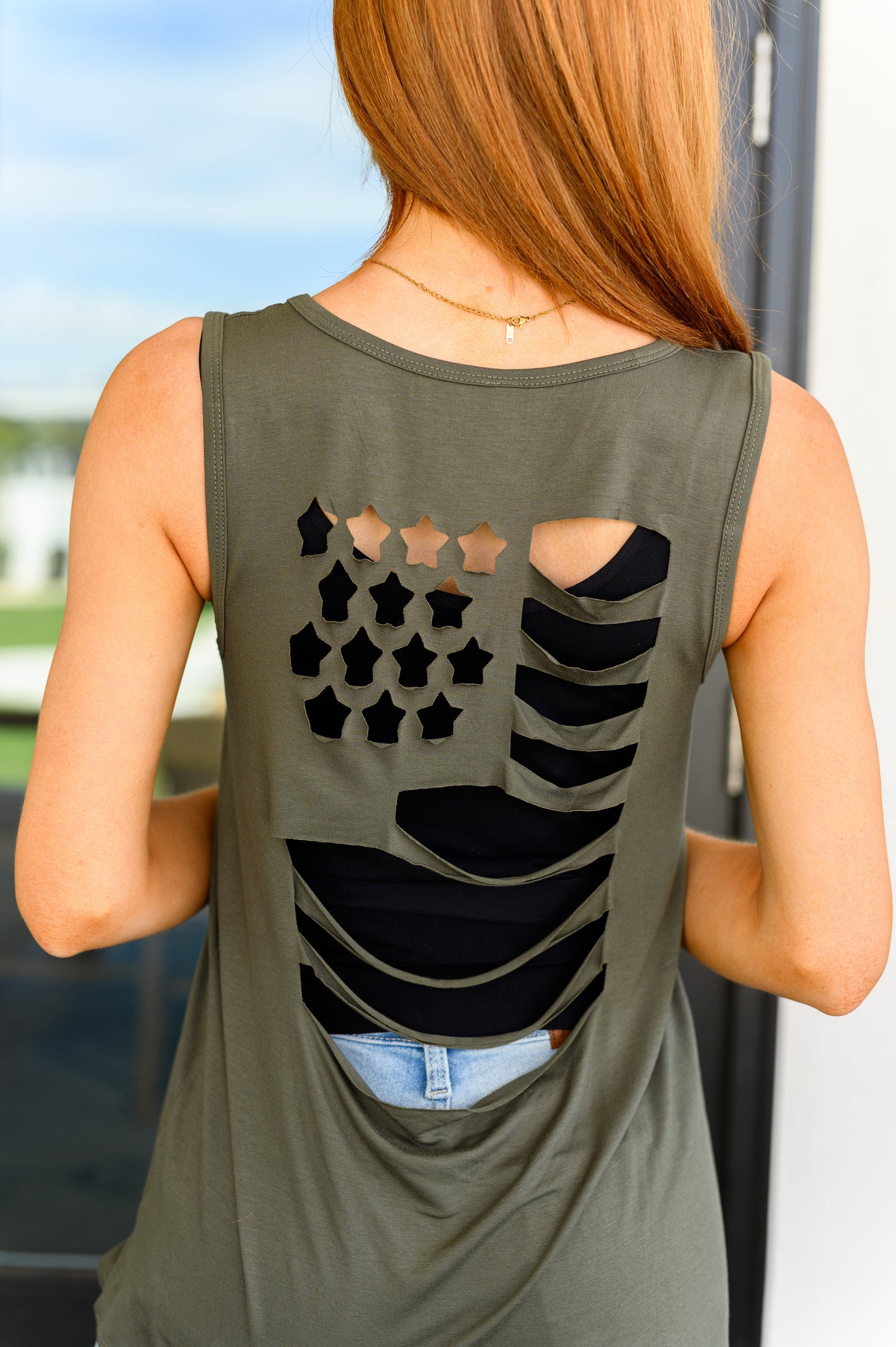 A woman wearing an olive green tank top with a laser cut flag design on the back, in a v-neckline cut.