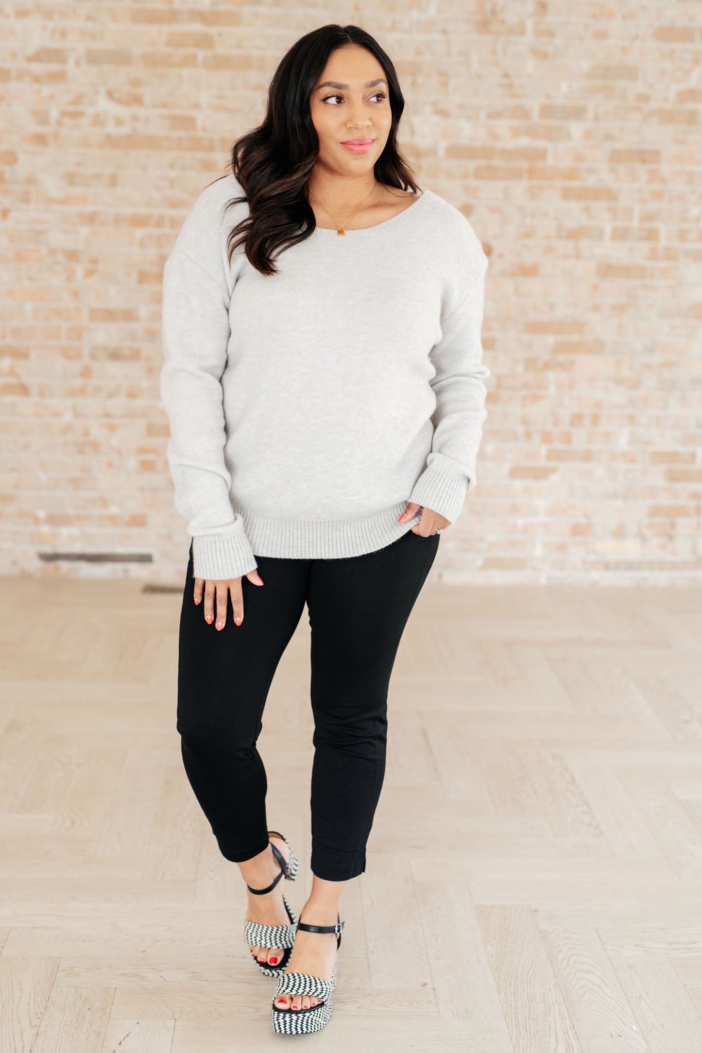 Woman wearing a light gray sweater and black pants against a brick wall.